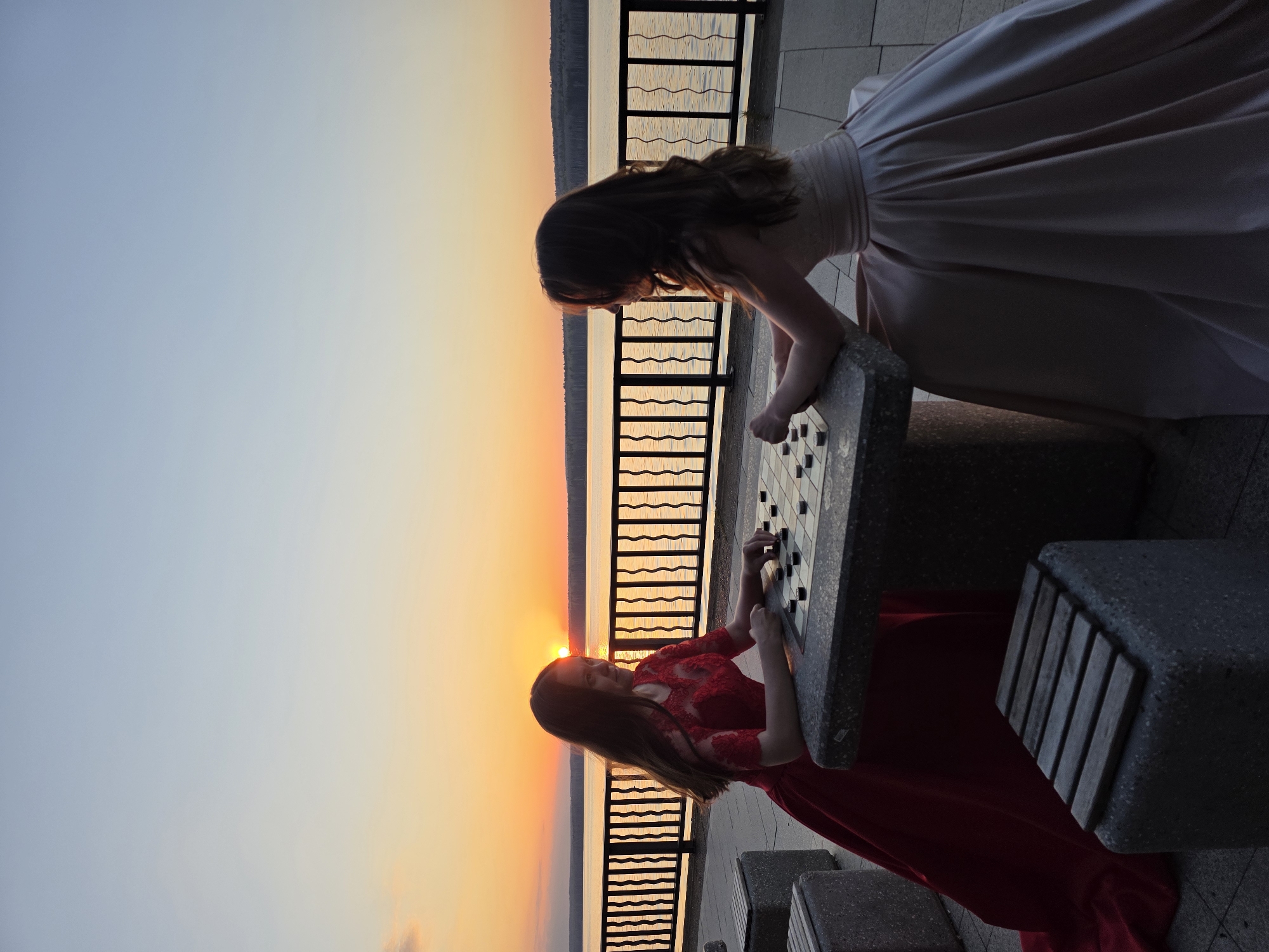 Two girls in evening gowns play checkers at a stone table along a waterfront promenade. The girl on the left wears a red gown, while the one on the right wears a blush-colored dress. A black metal railing separates the promenade from the water, where the sun is setting on the horizon. The scene is the embankment in the city of Chaikovsky, Perm Krai, Russia.