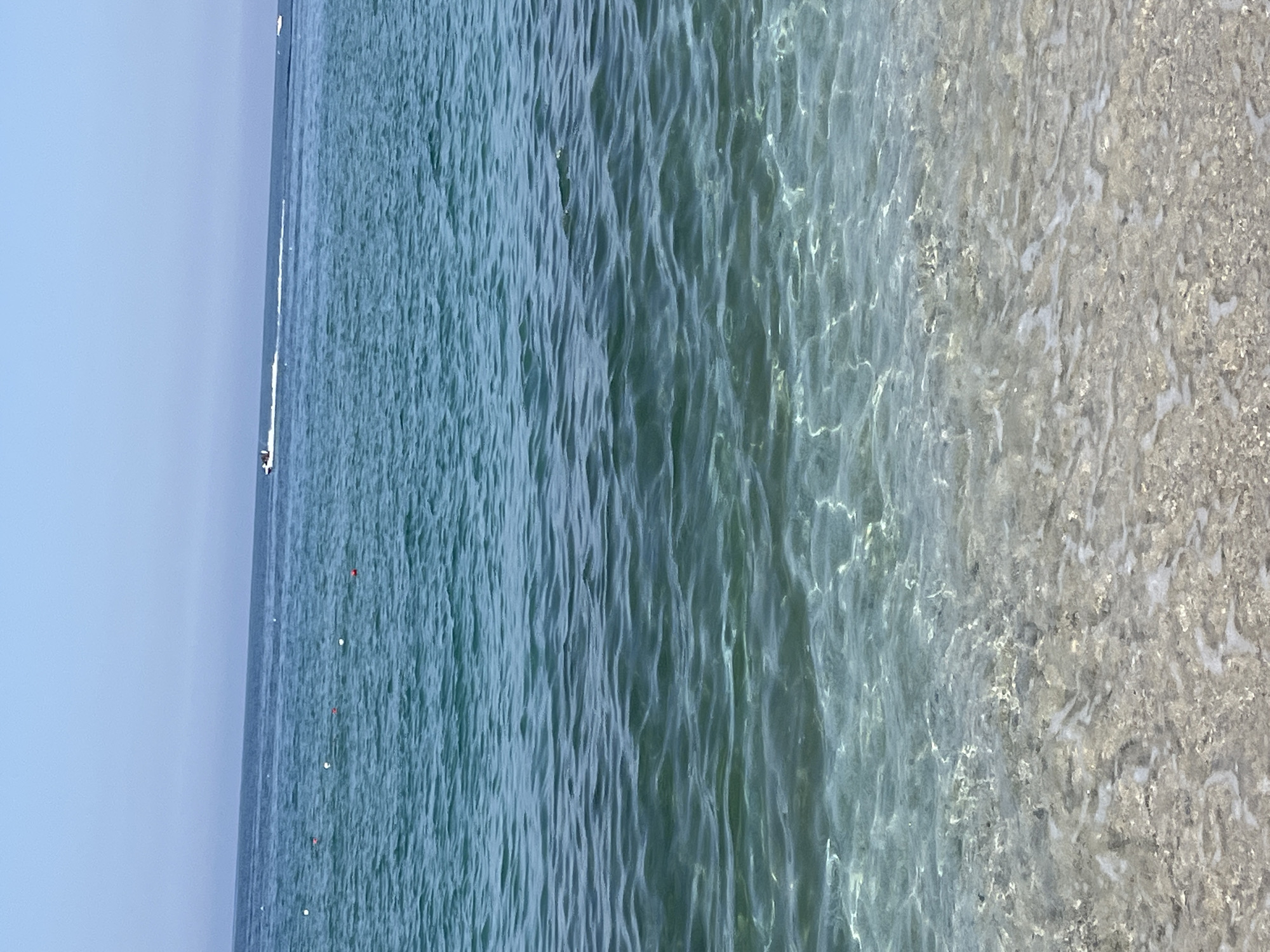 The image shows a beach scene at the Mediterranean Sea in Alanya, Turkey. The water transitions from a deeper blue-green hue in the distance to a clear, transparent color near the shore. Ripples are visible on the water surface, suggesting gentle waves or movement. A light-colored, sandy or pebbly beach is seen along the right edge. Small red and white floating objects are visible further offshore. In the very distant left is a ship. The sky is a clear, pale blue.