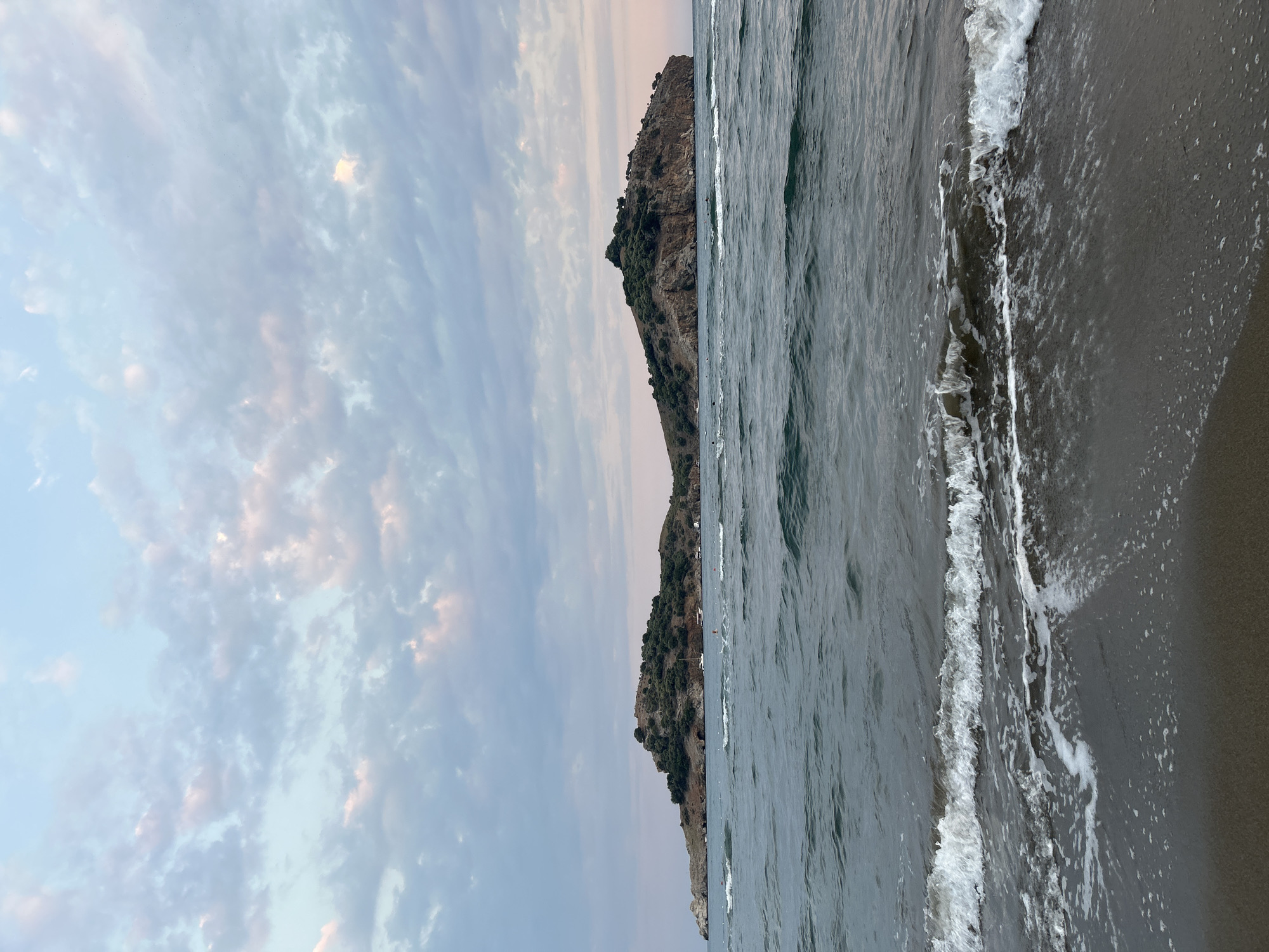The image shows a body of water with waves breaking on a dark sand beach in the foreground. In the mid-ground, there is a large, rocky island covered with trees. Several boats, including a white sailboat, are anchored near the island. The background consists of a sky with scattered clouds, tinged with pastel pink and blue hues, suggesting either dawn or dusk. The island is likely Pigeon Island (Güvercin Ada) in the Gulf of Gökova, near the town of Ortaca, Turkey. The scene depicts a tranquil coastal landscape.