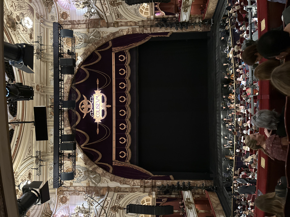 The image captures an interior view of a grand and ornate theater, seen from an elevated position in the audience looking towards the stage. The scene is dominated by a large, dark stage, currently empty, framed by a richly decorated proscenium arch and a heavy, dark purple curtain, possibly velvet. The curtain is adorned with elaborate gold trim, featuring a Greek key pattern, garland motifs, and circular laurel wreaths.
A prominent, illuminated sign hangs centrally above the stage, spelling out "COLISEUM" in stylized lettering, flanked by what appear to be golden eagle or phoenix figures. On both sides of the stage, large black loudspeakers and stage lights are mounted on visible scaffolding structures, suggesting a modern technical setup within the historic venue.
The theater's architecture is lavish, characterized by gilded moldings, decorative ceiling frescoes, chandeliers, and arched alcoves, indicative of a Baroque or Rococo style.
A significant audience is visible, occupying the stalls and boxes to the right of the stage (audience left). Most people are seated, with their backs or profiles towards the camera, looking towards the stage. A few individuals in the immediate foreground, including their heads and shoulders, are in view, emphasizing the high vantage point. The audience appears to be settled, waiting for or observing an event.
The artificial lighting suggests an evening event. The visible text "COLISEUM" directly identifies the venue, which, combined with the location context, confirms it as the London Coliseum in the United Kingdom.