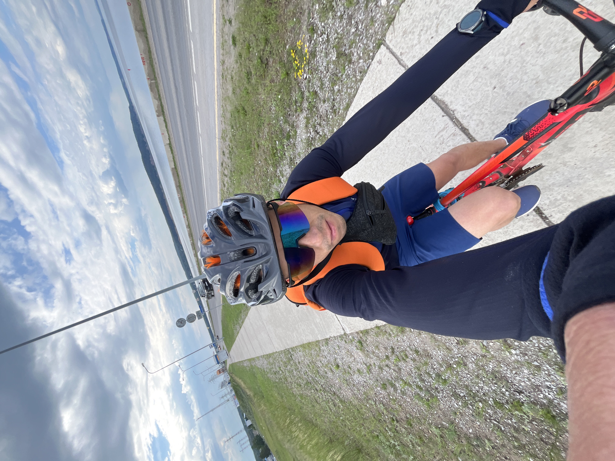 A male cyclist, wearing a grey and orange helmet, blue shorts, a dark blue long-sleeved shirt, and a bright orange vest, is riding a red bicycle on a concrete path alongside a road. He is also wearing reflective sunglasses and a dark waist pack. The cyclist is in motion, as indicated by the blurred image of the bicycle's frame and his arm extended forward, likely holding a camera or selfie stick.
The environment appears to be a semi-urban or suburban area, with a paved road and grassy verges. In the background, there is a body of water, possibly a reservoir or river, with a distant treeline. The sky is cloudy with patches of blue. The road is lined with streetlights and utility poles.
The location is identified as Chaikovsky, Russia. The scene depicts outdoor recreational activity, specifically cycling, with elements of infrastructure and natural landscape.