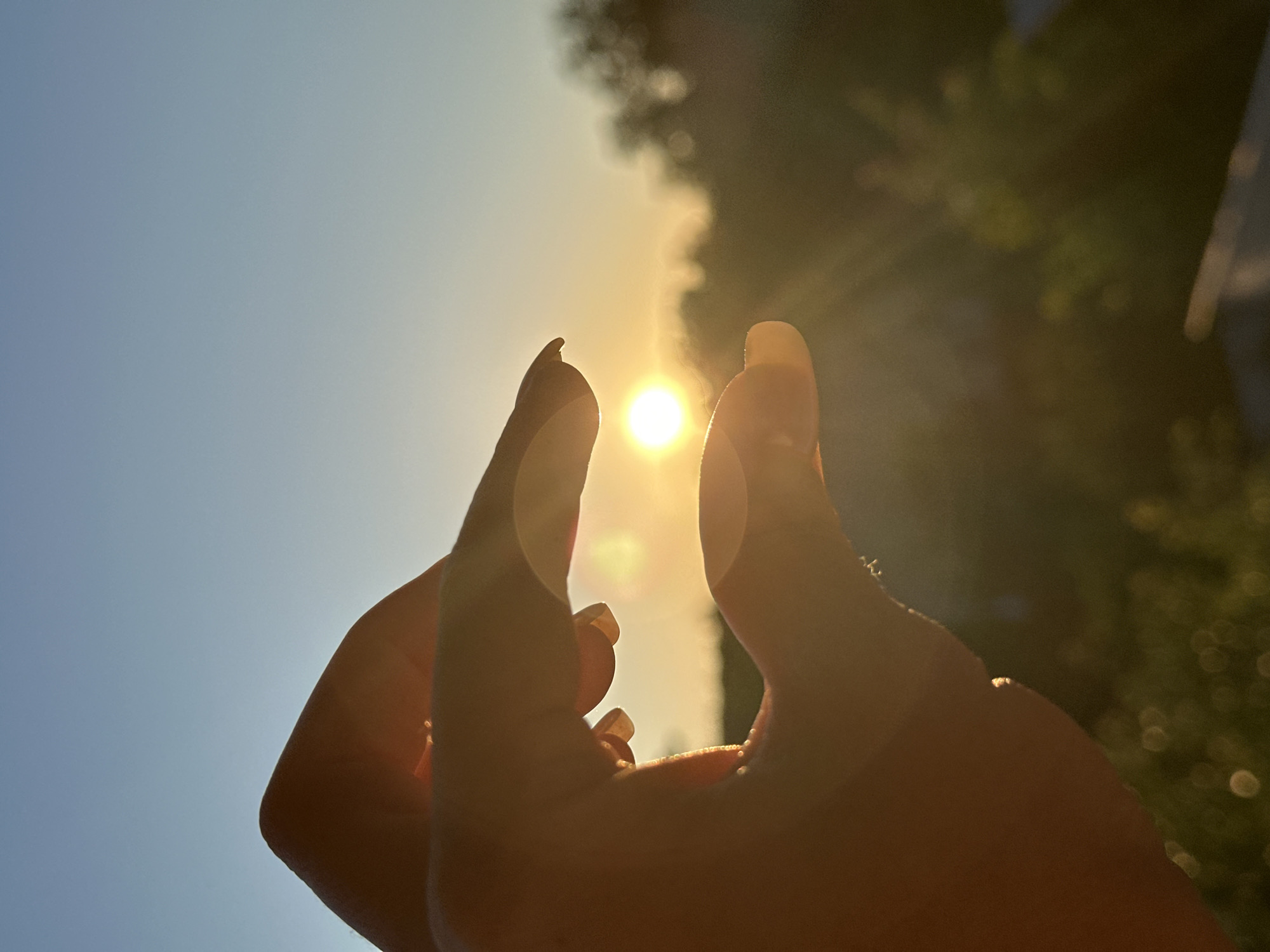A close-up, silhouetted shot captures a person's hands framing the setting sun. The sun appears as a bright, small orb of light, casting warm golden rays and lens flares across the image. The hands are cupped, with the thumb and index finger of each hand meeting, creating a small opening through which the sun is viewed. The background is blurred, showing the warm hues of the sky transitioning from orange near the horizon to a soft blue overhead. Silhouetted trees and foliage are visible in the middle ground, suggesting an outdoor setting. The image was taken at sunset in the city of Nizhny Novgorod, Russia.