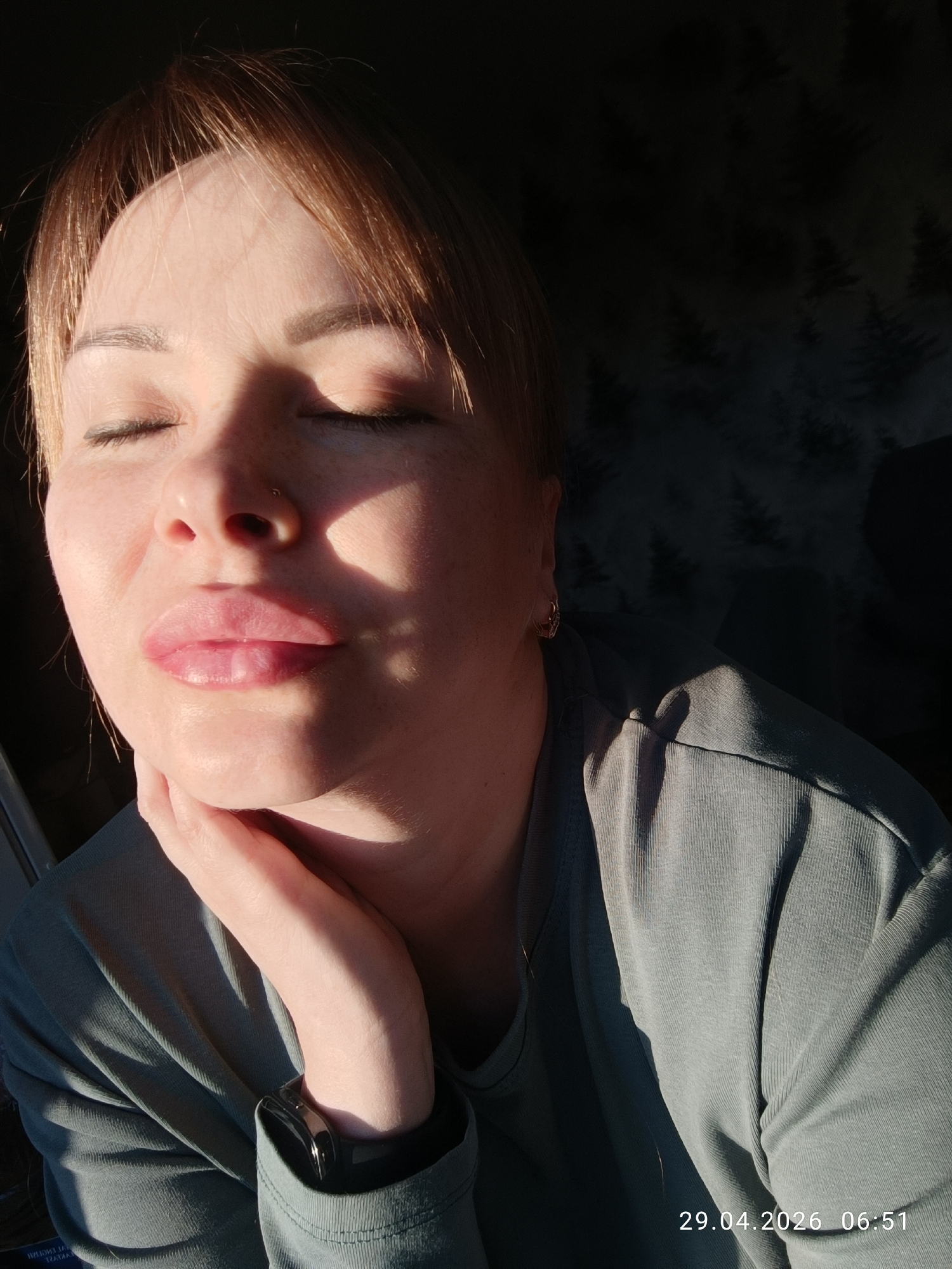 The image presents a close-up portrait of a woman, likely captured indoors in Chaikovsky, Russia, during the early morning. She is depicted with her eyes closed and lips softly pursed, her left hand resting gently under her chin, suggesting a moment of peace, relaxation, or playful enjoyment of the sunlight. Strong, golden sunlight illuminates the right side of her face, neck, and upper torso, creating a high-contrast scene with deep shadows on her left side and the background.
Her hair is light brown with a fringe, highlighted by the sun. Visible facial features include fair skin with some freckles, a small nose piercing, and defined eyebrows. She wears a muted grey-green ribbed long-sleeved top. A gold-colored hoop earring is visible in her right ear, and a dark smart watch is on her left wrist. The background is dark with a subtle, abstract pattern resembling silhouetted pine trees. In the bottom right corner, the text "29.04.2026 06:51" indicates the precise date and time the photo was taken. The strong, low-angle light suggests a clear and sunny start to the day.