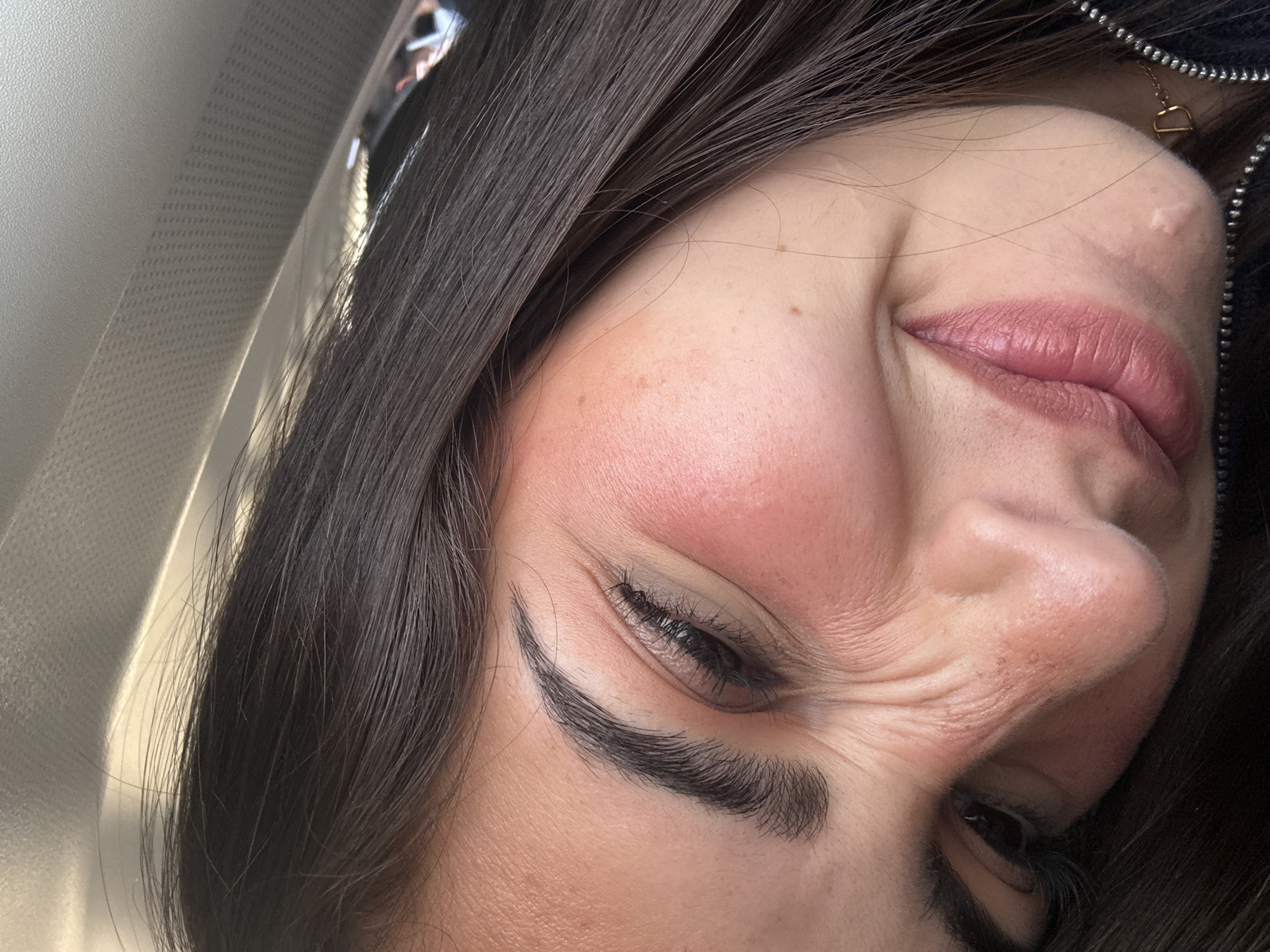 A close-up, slightly angled, portrait of a person with dark hair and fair skin. The person's right eye is almost fully closed, with the left eye slightly more open. Wrinkles are visible around the eyes and nose due to the facial expression. The lips are full and colored with a pinkish hue. The person appears to be inside a vehicle, as indicated by the material visible in the background, which resembles a car's interior. The image doesn't provide enough information to place the subject or scene in San Antonio, United States.