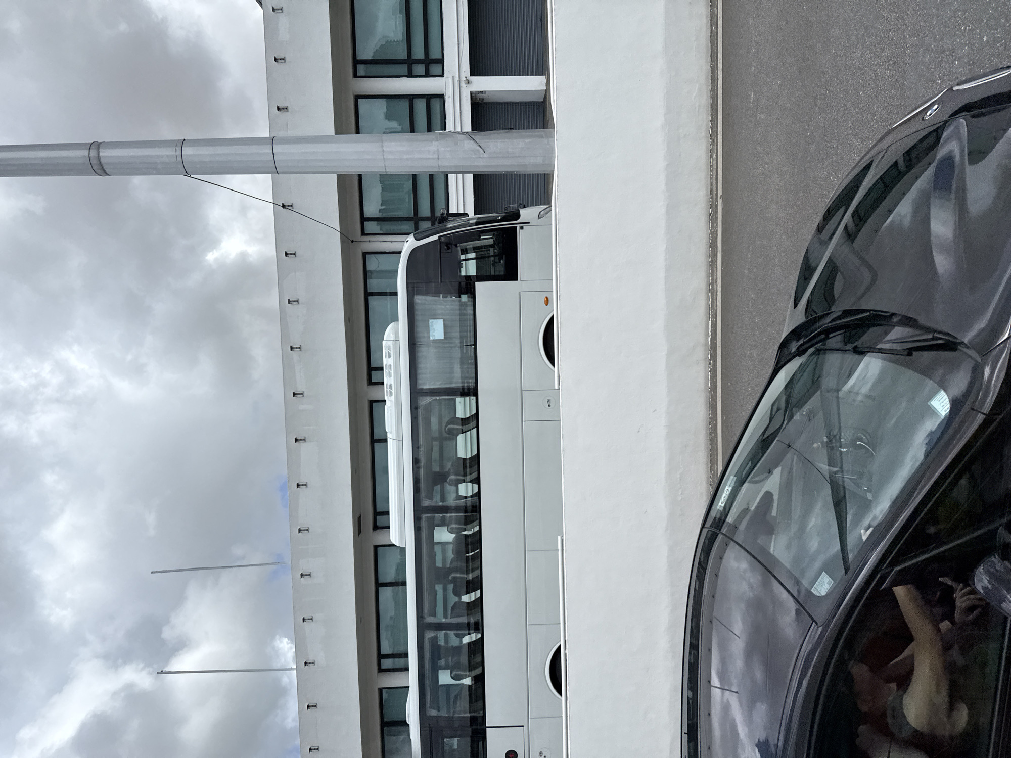 An exterior, eye-level, medium shot captures a white bus traveling past a modern building with large windows. The bus is partially obscured by a white concrete barrier in the foreground. A black car, also partially visible, is in the immediate foreground. The sky is overcast with heavy clouds. Two flagpoles stand on top of the building. The scene appears to be a street in Macau, China, likely near a transportation hub or commercial area.