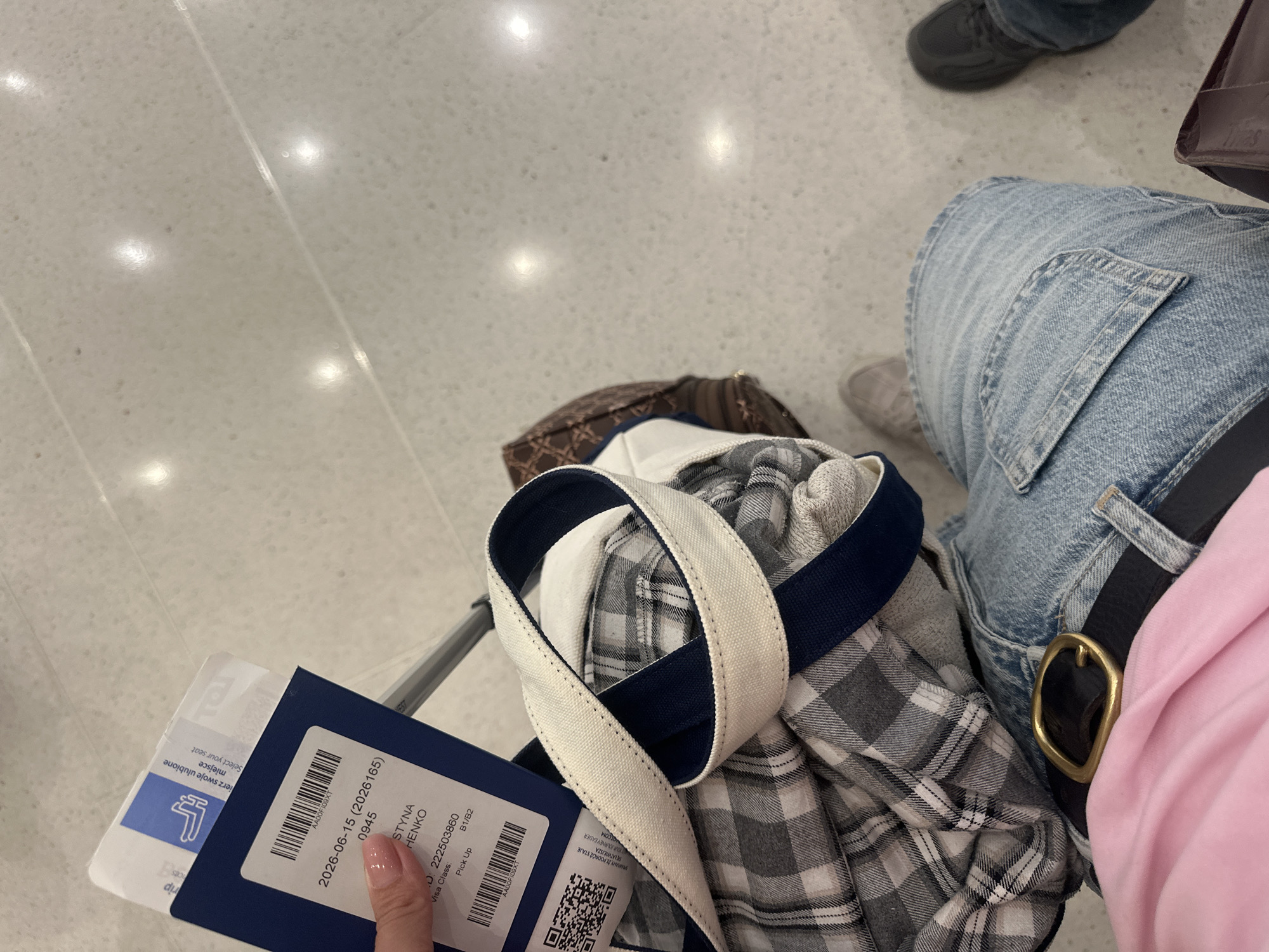 The image shows a person's legs and hands at an airport. The person is wearing blue jeans and a pink shirt. They are holding a boarding pass and a passport. The boarding pass has information about a flight, including the date, time, and passenger name. The passport is a blue booklet. In the background, there is a plaid shirt and a patterned bag. The floor is a light-colored, speckled surface, likely a common airport flooring material. The lighting suggests an indoor environment, possibly a gate area or waiting lounge. There are no clear indicators of the specific city or location within Chicago.