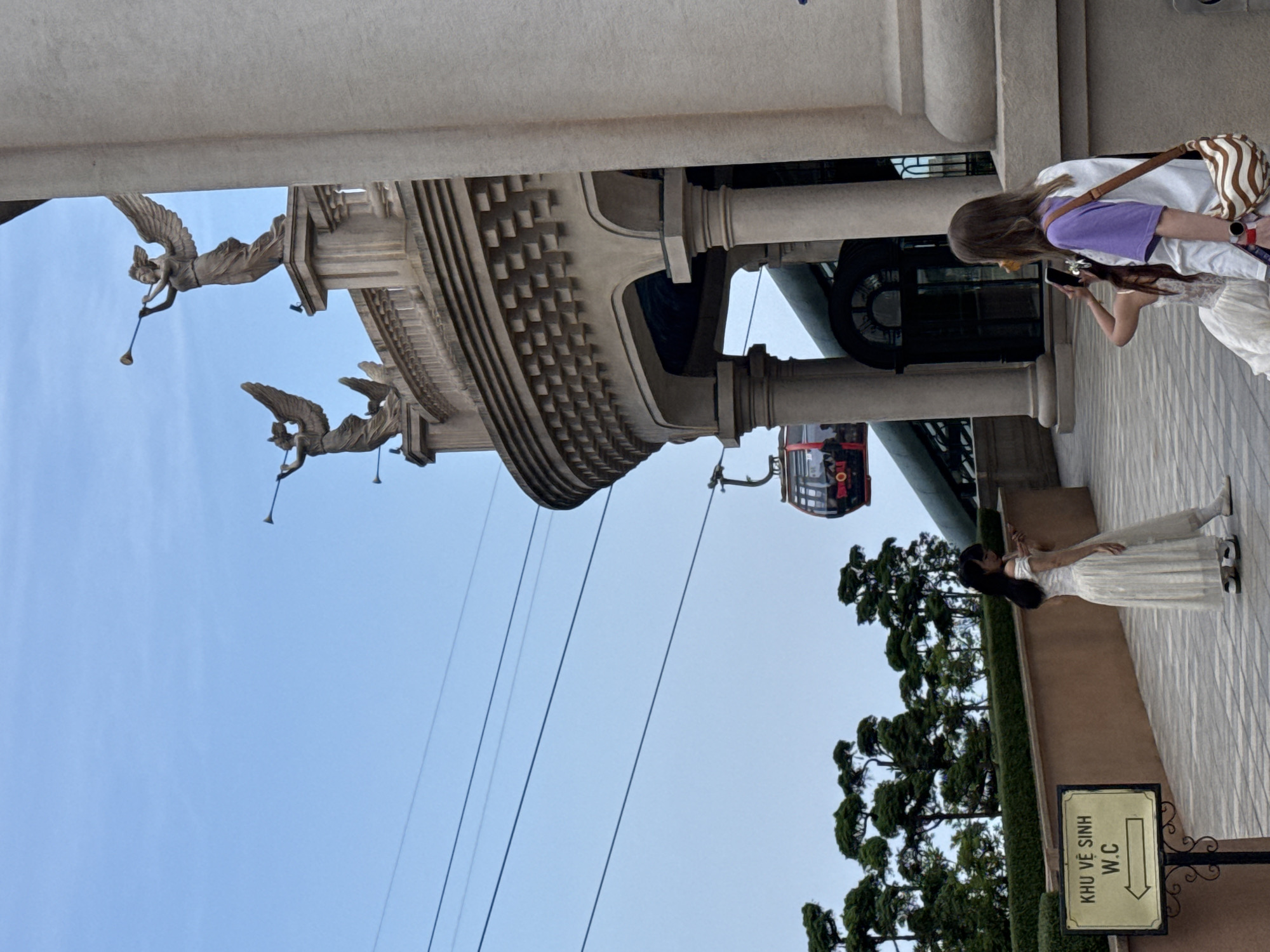 This image captures an outdoor scene at what appears to be a grand tourist attraction, possibly in Đà Nẵng, Vietnam, given the location context. The setting features ornate, classical-style architecture, a cable car system, and several individuals enjoying the environment.
The dominant subjects are a large building structure and two women. The building is elaborate, with columns, patterned cornices, and two large, winged sculptures resembling angels blowing trumpets, attached to its upper facade. Below, a red and black cable car gondola is visible, suspended on wires against the clear blue sky, suggesting an active transportation system.
In the foreground, a woman with long dark hair, dressed in a flowing white or light-colored gown, stands gracefully, her arms slightly extended, possibly posing for a photograph. Her posture conveys elegance and calm. Further back, another woman with long brown hair, wearing a purple top and a white outer layer, carries a striped shoulder bag. She is holding a smartphone, seemingly capturing a moment or an aspect of the surroundings. Another person's arm is visible next to her.
The overall activity taking place is sightseeing and enjoying the unique architecture and views. The time of day appears to be bright daytime, under a clear, cloudless blue sky. A notable detail is a signpost on the lower right, which reads "KHU VỆ SINH" (Vietnamese for "Restroom/Toilet area") and "W.C" with a right-pointing arrow, indicating facilities nearby. The elaborate sculptures and cable car system suggest a themed park or resort environment.