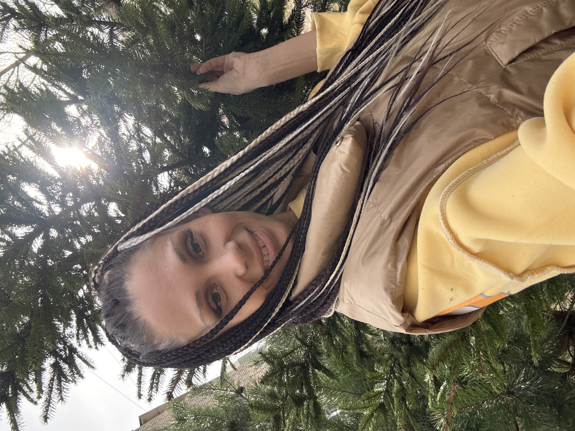 A low-angle shot shows a woman standing outside in what appears to be a residential area of Zhukovo, Russia. She wears a tan vest over a yellow sweatshirt and has long, dark hair with white and brown braids. She is smiling and looks toward the camera, with a section of coniferous tree branches visible behind and beside her. The sun is visible in the upper left of the frame, shining through the tree branches. Part of a building is visible in the distance, indicating an urban or suburban setting. The woman's hand is also touching a branch of the tree.