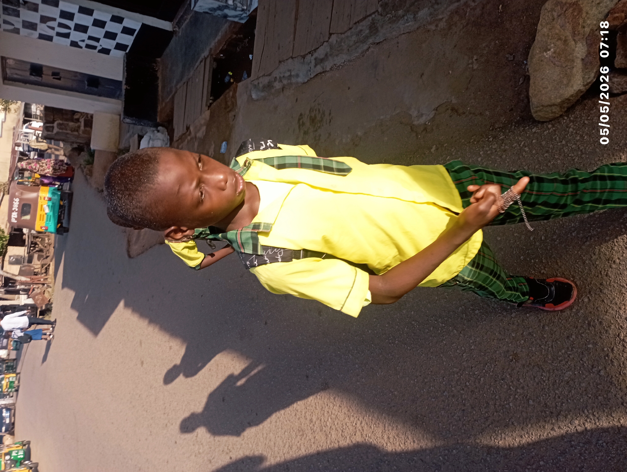 A young person stands on a paved street in Jos, Nigeria. The individual wears a yellow short-sleeved shirt with green and black plaid accents and matching plaid pants. He holds a silver-colored chain in one hand. Shadows of people and objects are cast on the street. In the background, there are buildings, vehicles (including auto-rickshaws), and people, suggesting an urban area. The date and time "05/05/2026 07:18" are visible in the image's upper-right corner.