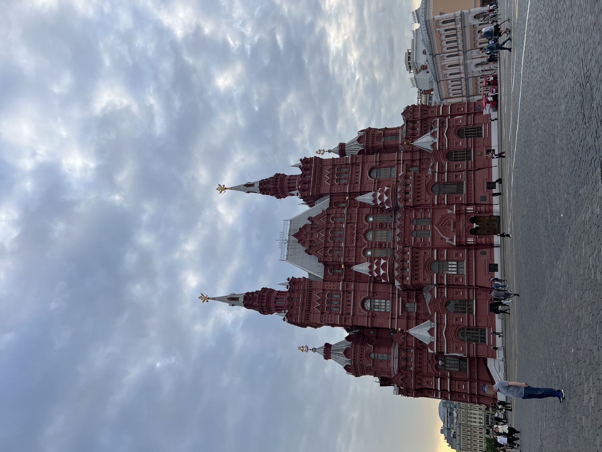 The image depicts the State Historical Museum located on Red Square in Moscow, Russia. The distinctive red brick building with its pointed towers stands prominently against a backdrop of a cloudy sky. People are walking around the square in front of the museum. The pavement appears to be cobblestone. Another building is partially visible to the right of the museum.