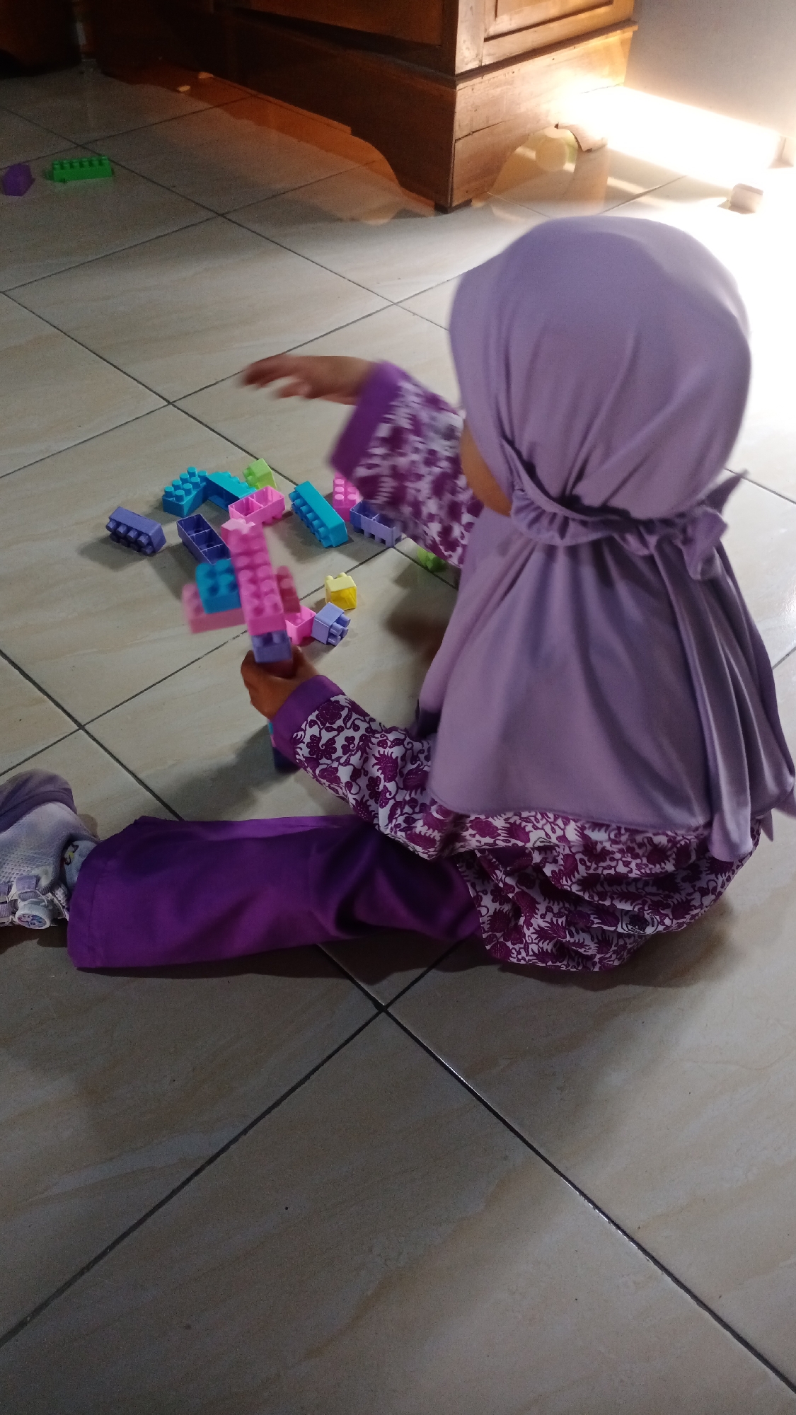 A young child wearing a purple hijab and a patterned top sits on a tiled floor, playing with colorful building blocks. The child is facing away from the camera, with their legs extended. The blocks are scattered around the child, with some in their hands. The floor is light-colored tile. In the background, a wooden cabinet is visible, and a bright light shines from the right side of the frame. The scene appears to be indoors. This image was taken in Mertan, Indonesia.