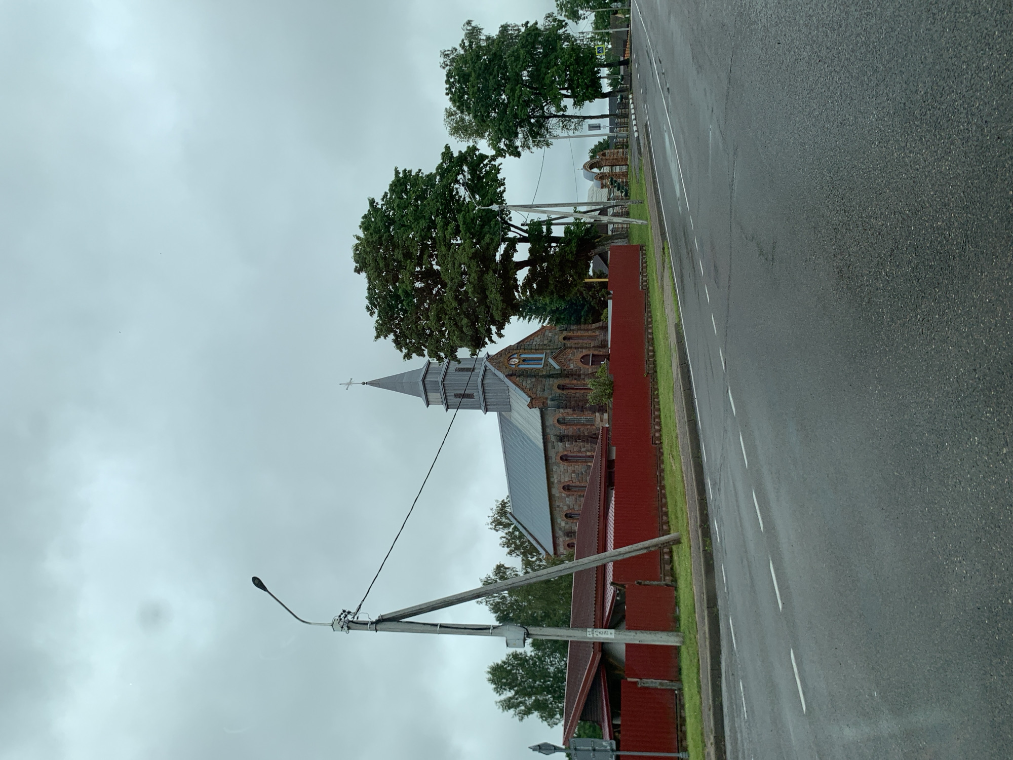 The image shows a view of a church and its surroundings on a cloudy day. In the foreground, a wet asphalt road with white dashed lines runs from left to right. To the left of the road, a bright red corrugated metal fence separates the road from a grassy area and a building with a red roof. Behind the fence, a stone church with a grey tiled roof and a tall steeple topped with a cross is visible. The church has arched windows and stonework. A large green tree stands next to the church. Further back, more trees and some other structures are visible. A street lamp is present on a pole to the left of the church. The sky is overcast and grey.