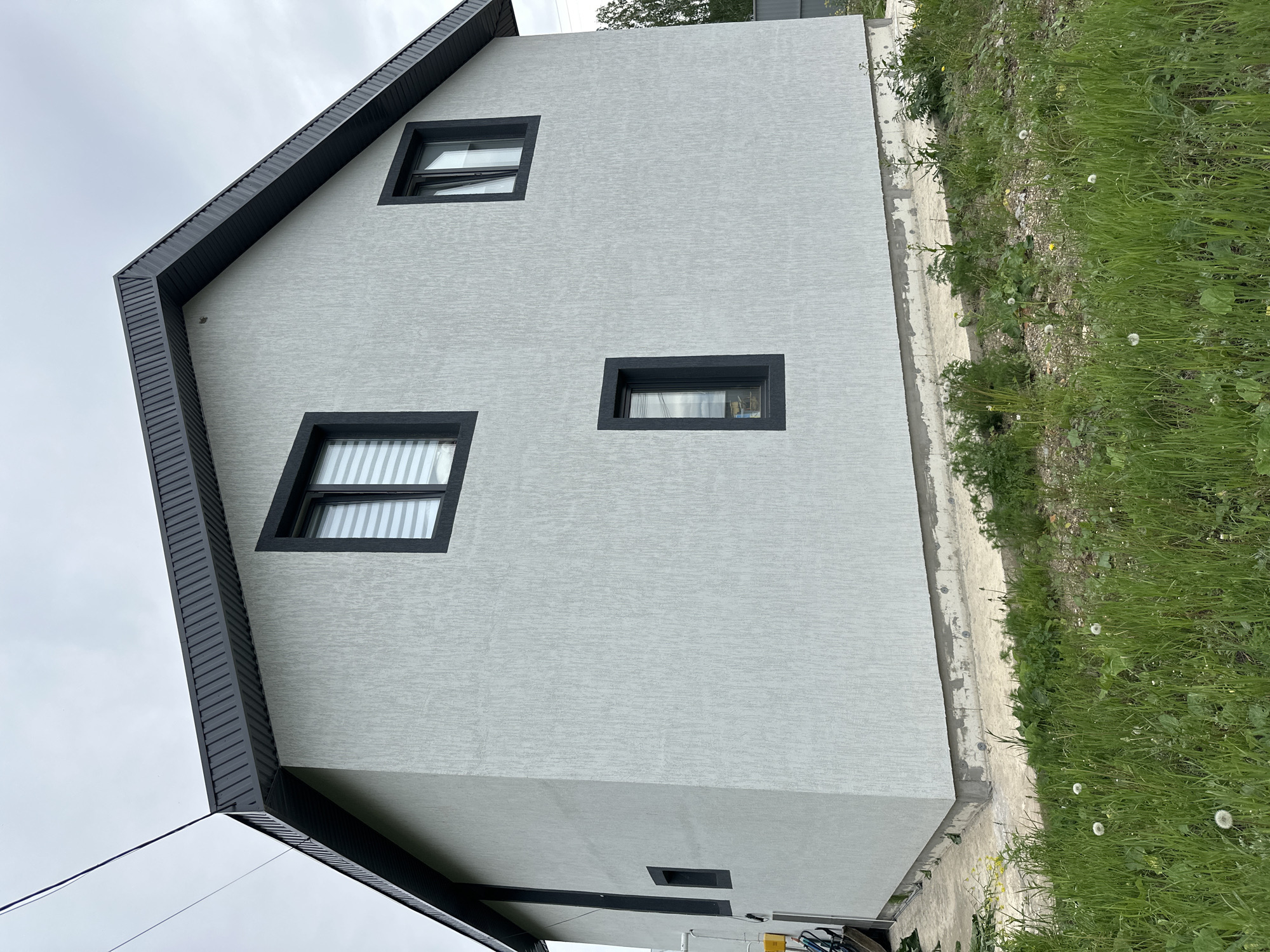 The image depicts a modern two-story house with a light gray stucco exterior and dark gray trim around the windows and roof. The house has a gable roof, and two visible windows on the upper story, and one smaller window visible on the first story. The windows are rectangular with black frames. The surrounding environment includes a patch of green grass with dandelions growing along the side of the house. The scene appears to be outdoors under an overcast sky. The location is identified as Unknown, Unknown.