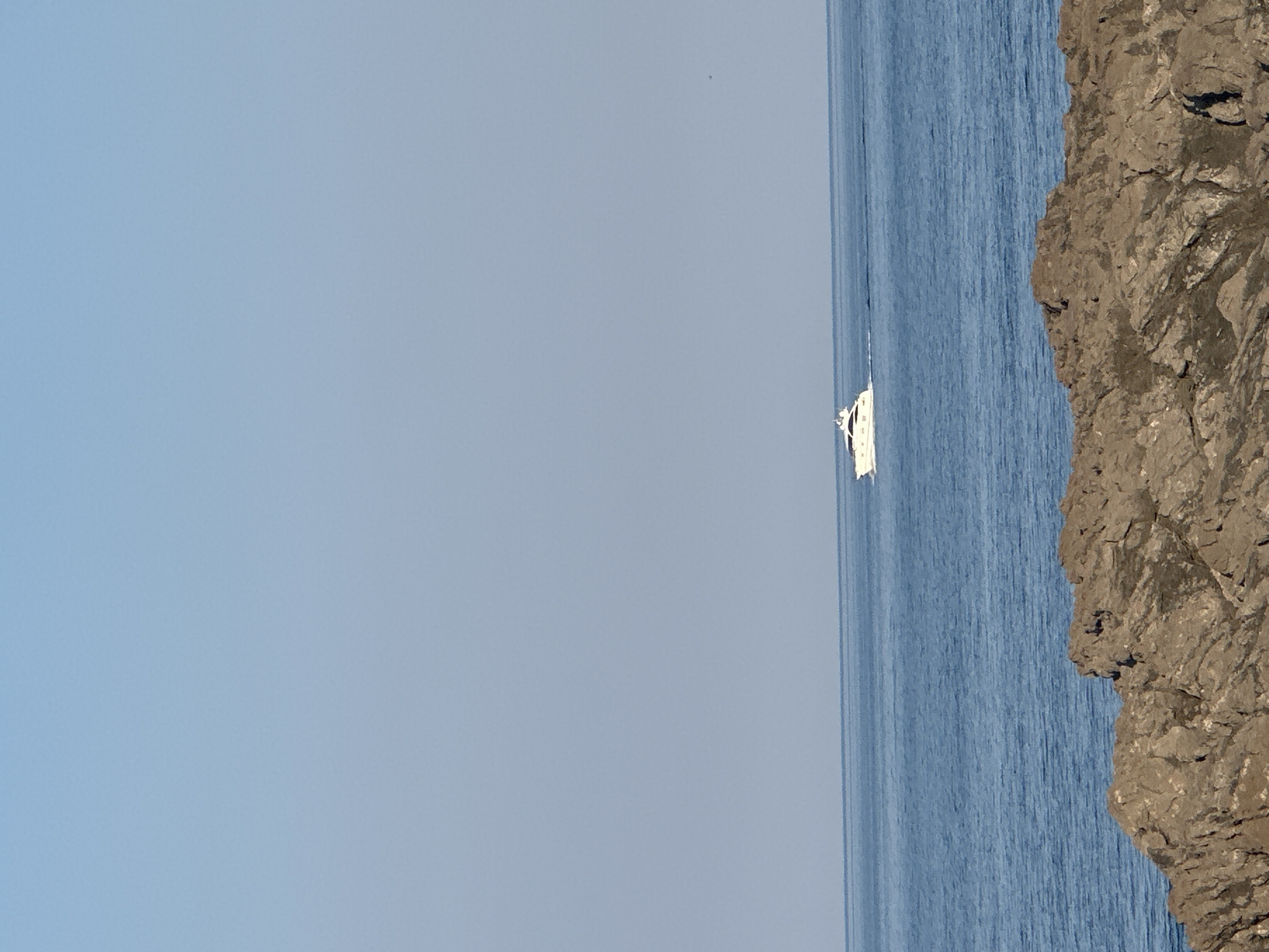 A white motor yacht is cruising on a calm, blue sea. The yacht is white with dark windows and a sleek design. It is leaving a white wake behind it as it moves from left to right across the frame. In the foreground, there are large, rough, brown rocks. The sky above is a clear, pale blue. The scene is likely near the coast, possibly in or around Kubasi, Montenegro, given the rocky shoreline and the presence of the sea. There are no people visible in the image.