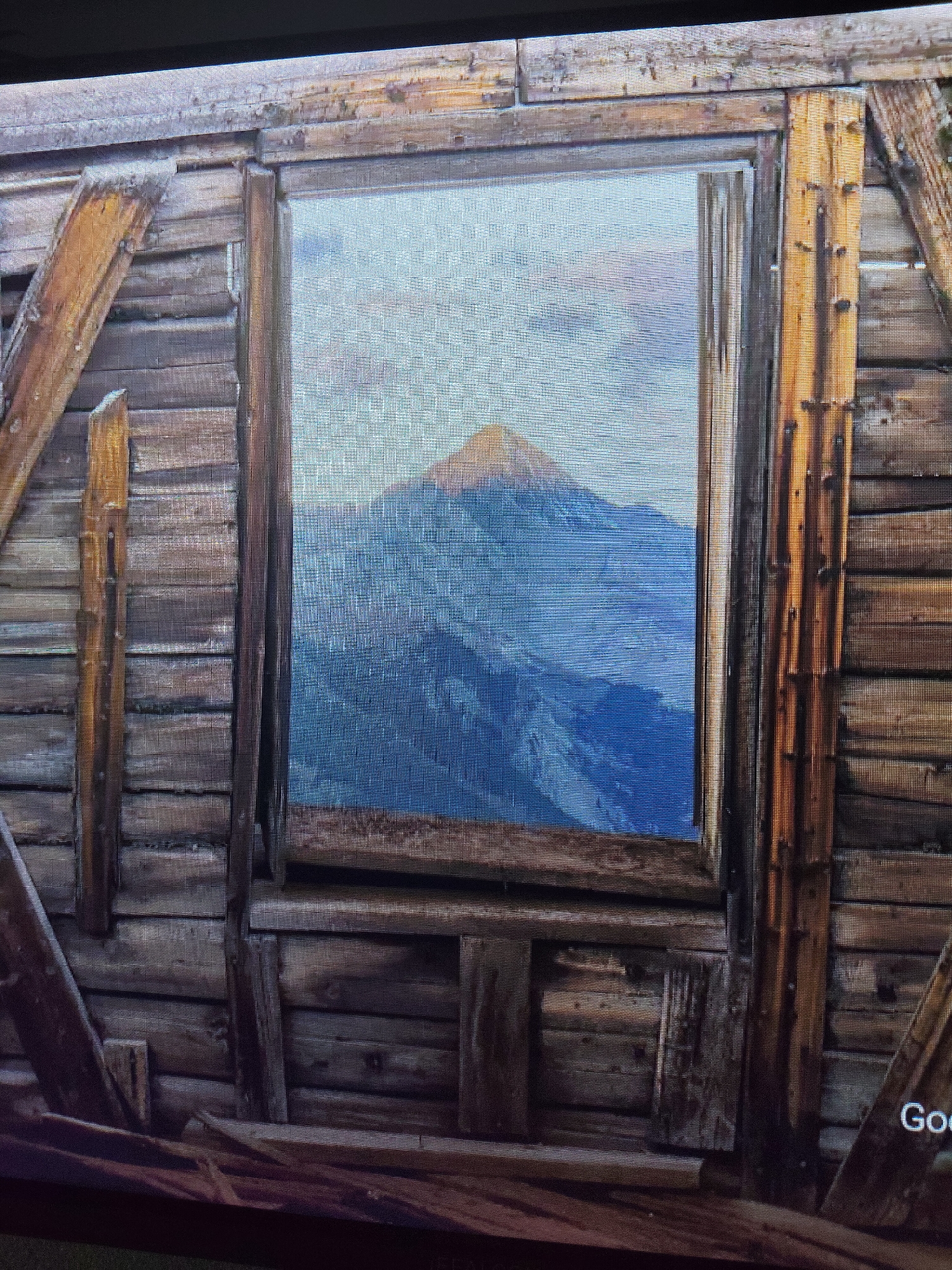 The image shows a close-up of a rustic wooden structure with a window. Through the window, a mountain landscape is visible, with a peak in the center under a cloudy sky. The wooden walls appear weathered and are constructed from horizontal planks. Diagonal wooden supports are attached to the exterior. The bottom right corner of the image shows text that appears to be a watermark or logo, with the word "Goo" visible. There are no people or animals in the image. The scene is not identifiable as being in Reftinsky, Russia.