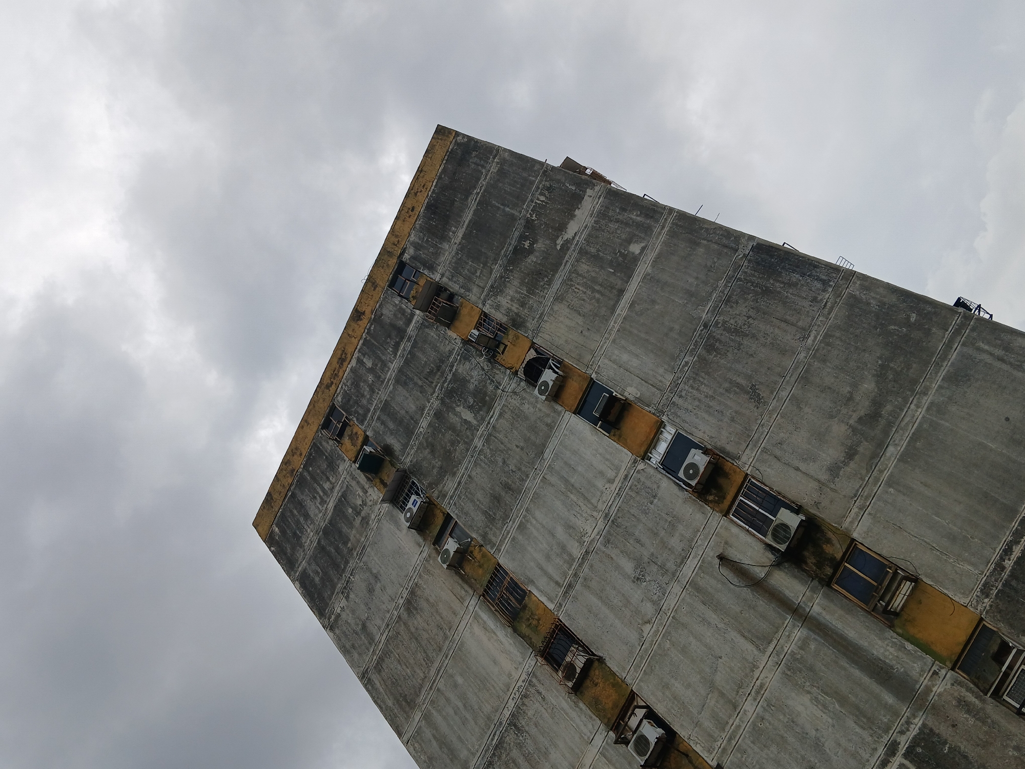 An upward-angled view shows a tall, rectangular concrete building against a cloudy sky. The building has a repetitive pattern of vertically oriented concrete panels, with a row of window openings between each set of panels. Air conditioning units are affixed to some of the window openings. The edges of the building are marked with a painted yellow stripe. The building's concrete is weathered and stained with gray marks. The scene is located in Ciudad Guayana, Venezuela.