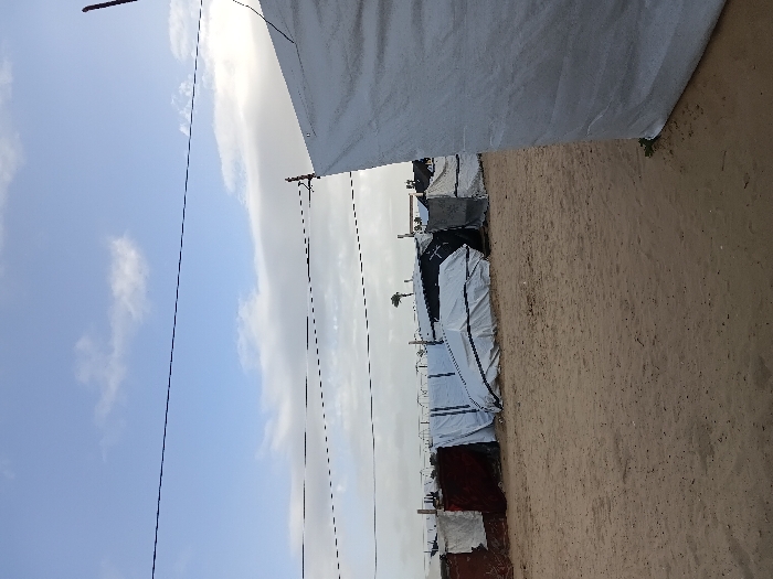 A line of tents stretches into the distance under a cloudy sky. The tents are primarily white, some appearing darker, and are pitched on sandy ground. Overhead power lines crisscross the sky. The view is obscured by a large, white structure in the upper portion of the frame.