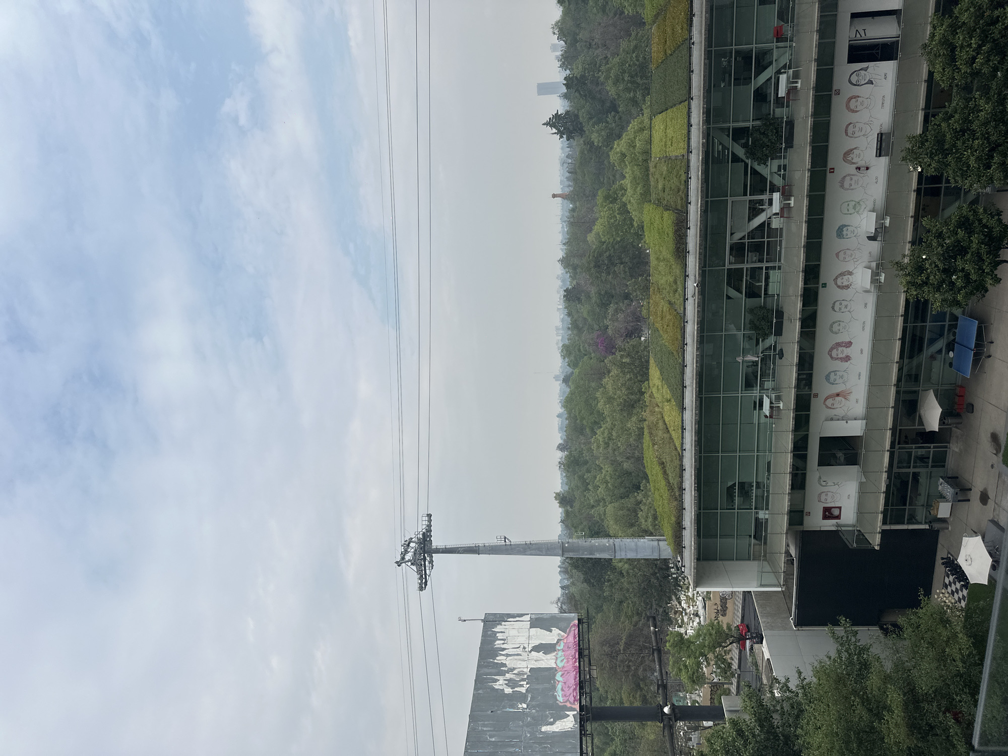 The image shows a view in Mexico City, Mexico. The sky is overcast with light blue and grey clouds. In the foreground is a cable car pylon and overhead wires. A large, weathered billboard is also visible, partially obscuring a view of the green trees.
To the right, a modern building with a glass facade is visible. It has a green living wall and banners with sketched portraits on its exterior. A cafe is on the street level.
In the distance is a skyline obscured by hazy conditions and vegetation.