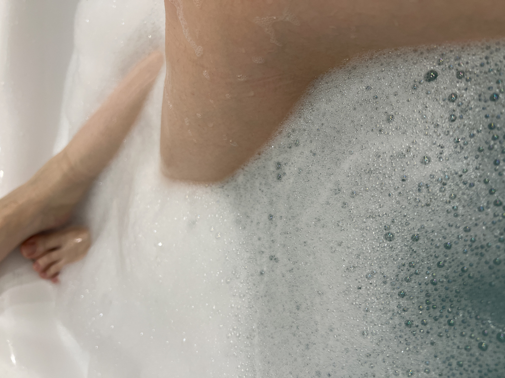 The image shows a partial view of a person's legs and feet submerged in a bathtub filled with soapy water and bubbles. The scene appears to be a close-up shot taken from above. The location in the city of Tyumen, Russia, cannot be determined from the visual information.