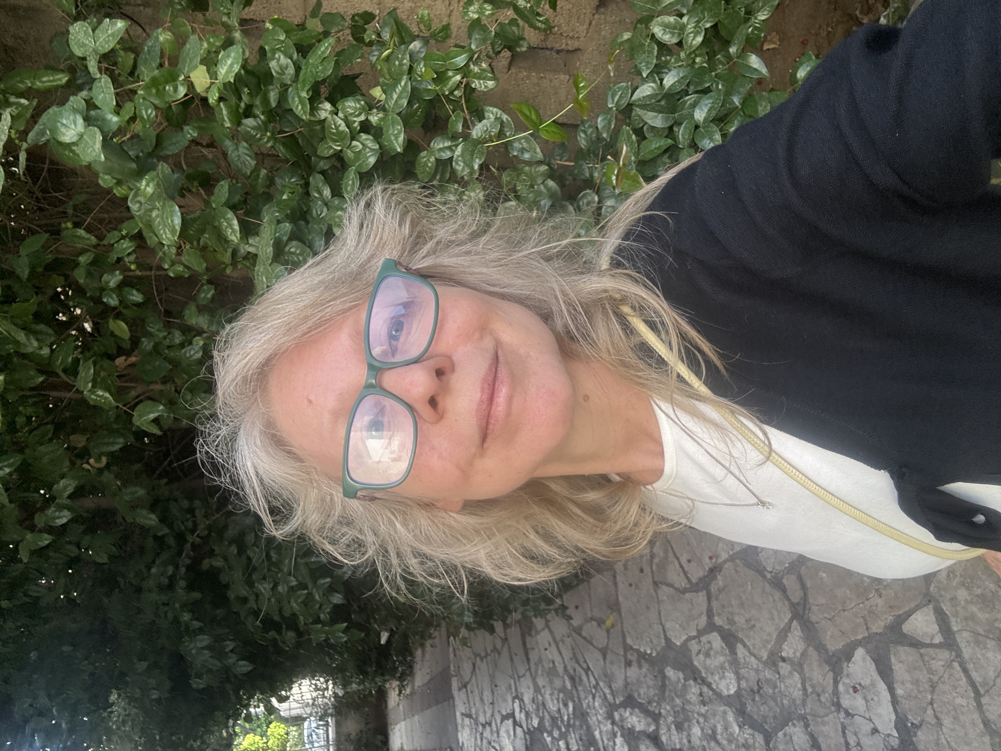 A woman is posing for a selfie outdoors in Ramat Gan, Israel. She has shoulder-length, graying blonde hair and is wearing green-framed glasses, a white T-shirt, and a black cardigan. A thin, gold-colored cord is draped around her neck. Behind her is a dense green wall of foliage. The ground she is standing on is paved with light gray stone slabs with dark gray grout. The location is urban.