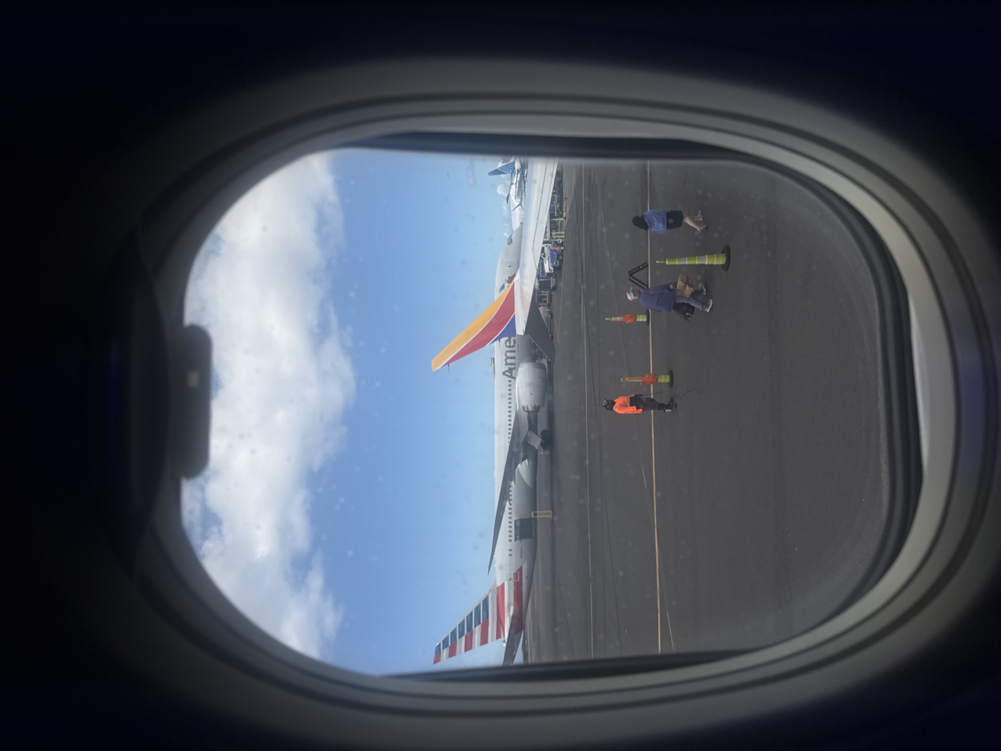 The image, captured through an aircraft window exhibiting minor surface smudges, depicts an airport tarmac under a bright, partly cloudy blue sky. A large American Airlines Boeing 787-9 aircraft, with the partial registration "N819AN" visible on its fuselage, is centrally parked, showcasing its distinctive red, white, and blue striped tail fin. To its right, partially obscured, the yellow, orange, and red striped tail of a Southwest Airlines aircraft is visible, with its wing extending towards the foreground. In the background, a blue tail fin of a third aircraft is discernible. On the dark asphalt tarmac, a single ground crew member wearing an orange high-visibility shirt and dark trousers stands to the left. Two individuals, likely passengers, walk from left to right across the tarmac; one wears a blue jacket and carries a dark bag, while the other wears a light-colored hat, a purple top, and carries several light-brown paper bags. Multiple green-and-orange traffic cones are positioned on the tarmac, delineating a path. Various pieces of ground support equipment are visible between the two main aircraft. This scene is located at Daniel K. Inouye International Airport (HNL), serving the city of Kailua, United States of America.