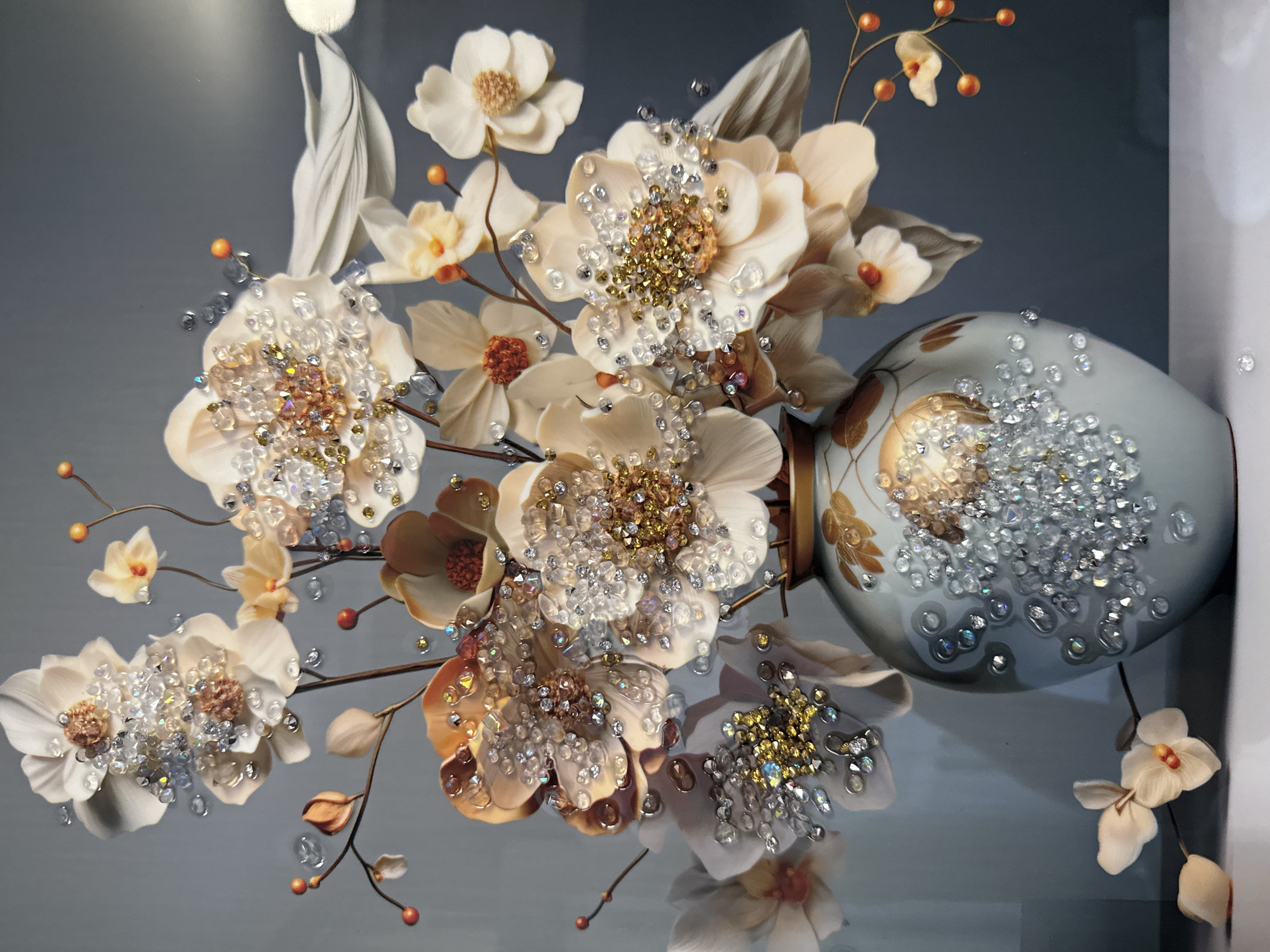 The image displays a close-up of a decorative vase filled with elaborate artificial flowers. The vase is light blue with gold floral accents and is heavily adorned with clear and gold rhinestones. The flowers themselves are also embellished with numerous rhinestones, creating a sparkling effect. Some flowers are white and tan, while others are a soft peach color. Small orange berries and thin brown stems are interspersed among the flowers. The background is a solid, muted gray. The lighting suggests the scene is indoors, possibly in a studio setting. There is no indication of the city of Tbilisi, Georgia, or any specific outdoor environment.