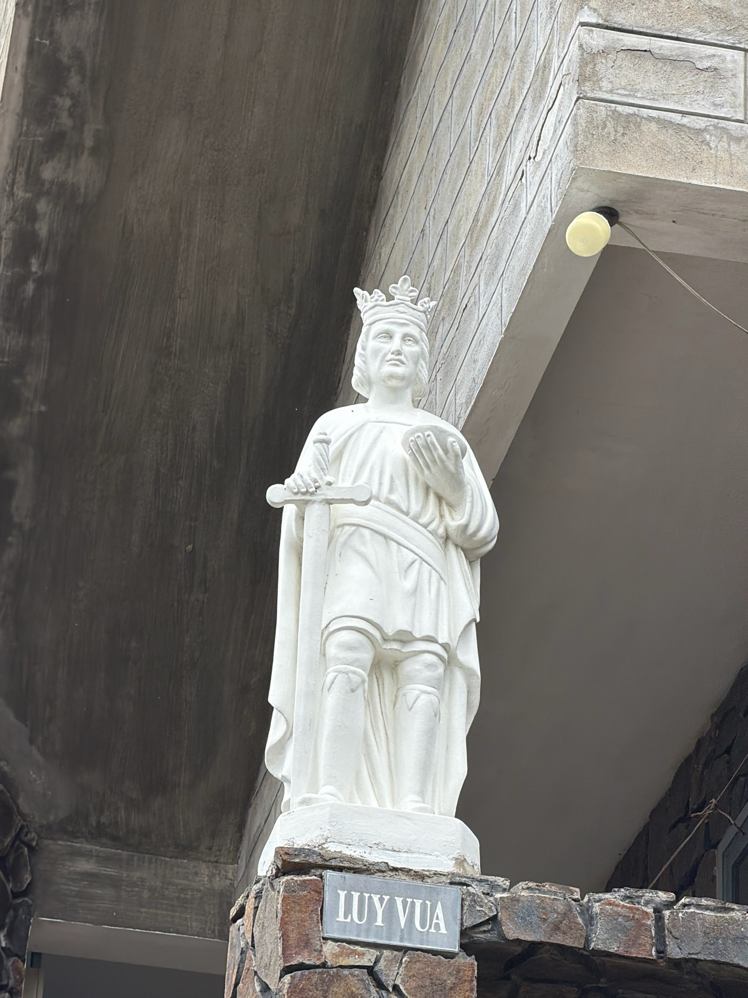 This is a ground-level, medium shot of a white statue of a king or a prominent historical figure, possibly Saint Louis or Ly The Vuong, in Nha Trang, Vietnam. The statue is made of white stone or plaster and depicts a male figure wearing a crown, holding a sword in his left hand and a round object in his right. He is dressed in flowing robes and armor on his legs. The statue stands on a base of rough-hewn stones, with a metal plaque at the bottom reading "LUY VUA". The statue is positioned in front of a building with a textured, grey concrete overhang and a wall constructed of light-colored bricks. A yellow bulb light fixture is visible attached to the underside of the overhang. The overall setting suggests an outdoor architectural element, possibly part of a church, monument, or public building.