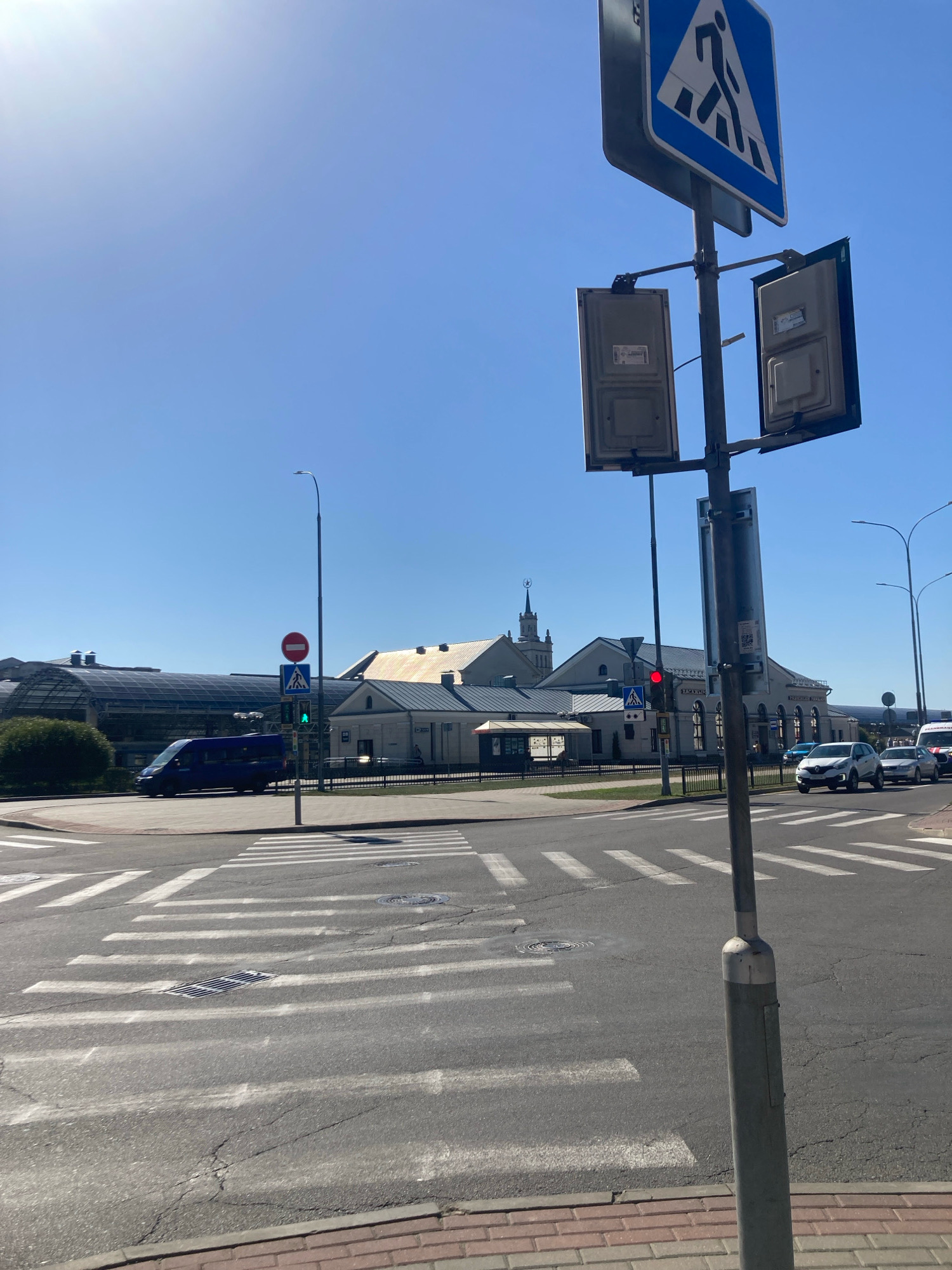 The image depicts a sunny daytime scene at a crosswalk near a train station in Brest, Belarus. A blue van is visible on the road, and several cars are stopped or moving slowly. Pedestrian crossing signs and traffic lights are present. The architecture of the train station is characterized by a long, white building with a yellowish roof, and a modern glass-covered structure adjacent to it. The sky is clear and blue. The foreground shows a paved pedestrian area with red bricks, leading to the crosswalk marked with white stripes on the asphalt. A streetlight with attached electrical boxes stands prominently in the right foreground. The scene appears to be a typical urban environment with moderate traffic.