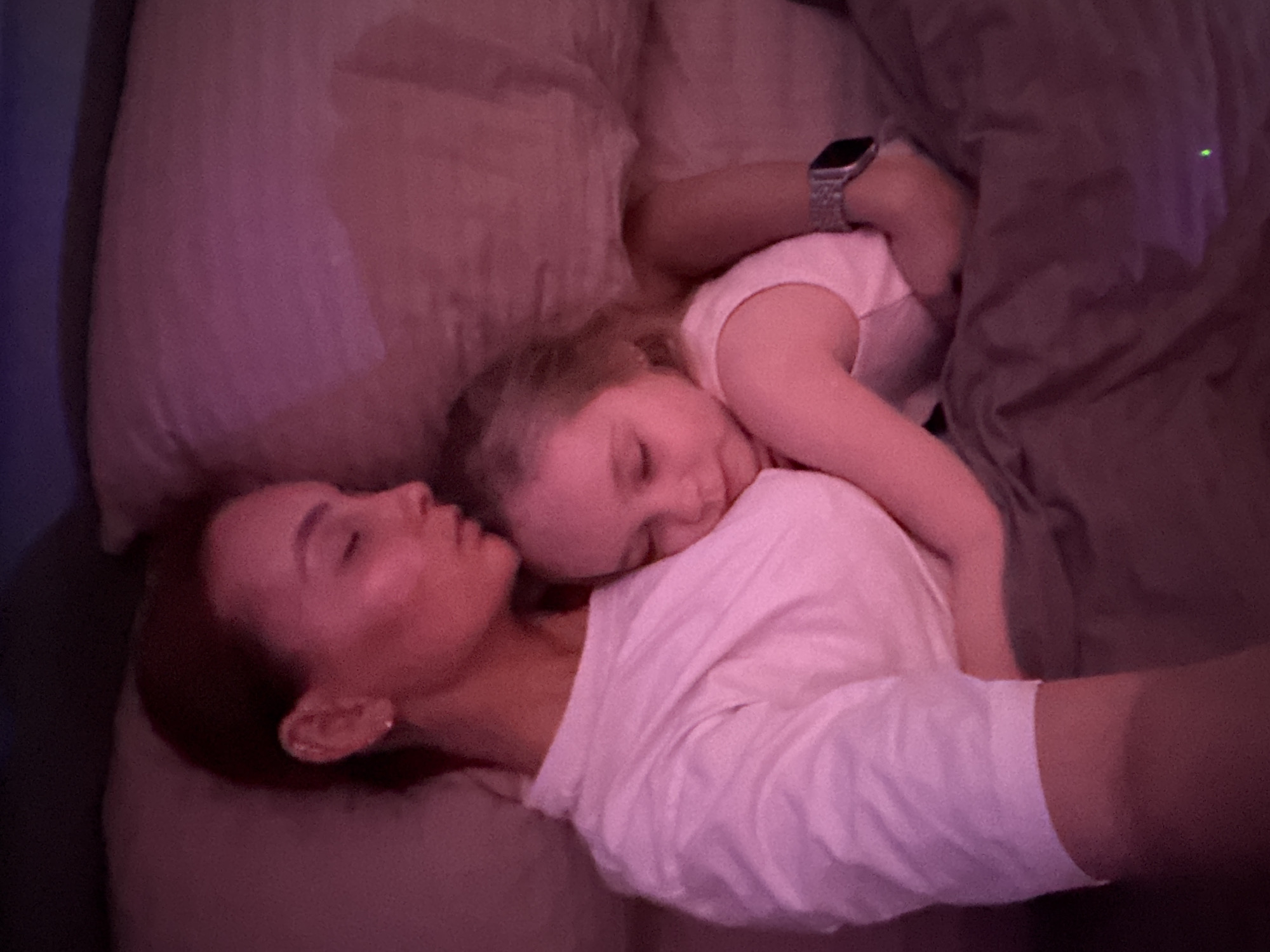 A medium shot depicts an adult and child lying on a bed. The adult lies on their back, face turned to the left, with the child lying on the adult's chest, facing the same direction. Both are sleeping. The adult has short, dark hair, and wears a white t-shirt. The child has blonde hair. A silver smartwatch is visible on the child's arm. The scene is dimly lit. The background is indistinct but includes pillows and a dark blanket. There is no verifiable information about the location being the city of Chaikovsky, Russia, in the provided image.