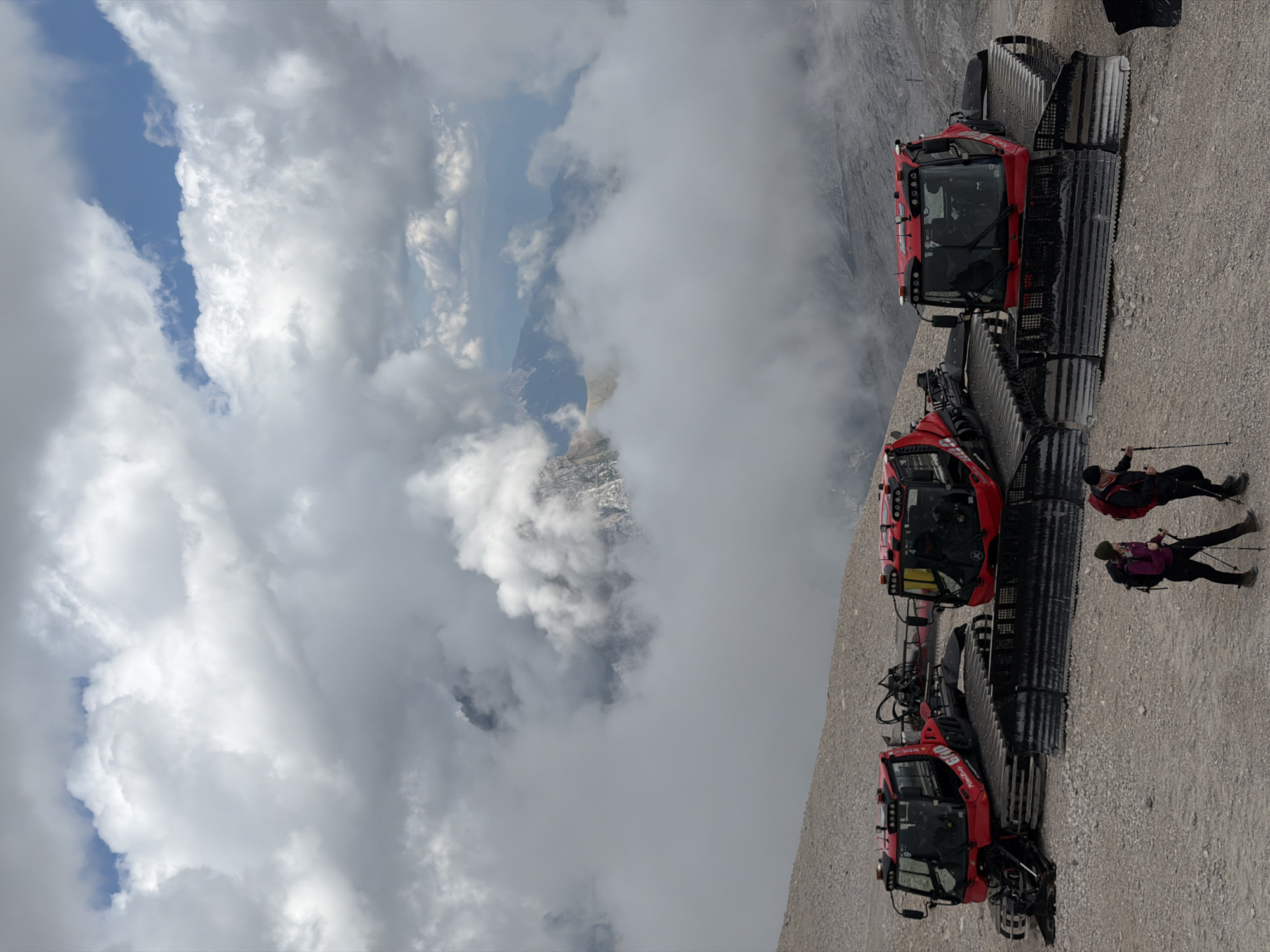 The image shows a steep, gravelly slope with three red snowcats parked in a line. Two individuals are visible near the bottom of the image, one standing with trekking poles and the other leaning on one. They are wearing outdoor clothing and backpacks. The snowcats are large, tracked vehicles designed for snow grooming or transport. The background features a dramatic sky filled with white and gray clouds, with glimpses of a valley below through breaks in the clouds. The scene suggests a mountainous environment, possibly during daylight hours given the brightness of the sky and the lighting on the snowcats. The location context provided is Garmisch-Partenkirchen, Germany, which is known for its alpine scenery and winter sports. Text is visible on the snowcats, including branding and what appears to be a model number or identifier. The overall impression is one of adventure or exploration in a rugged, high-altitude setting.