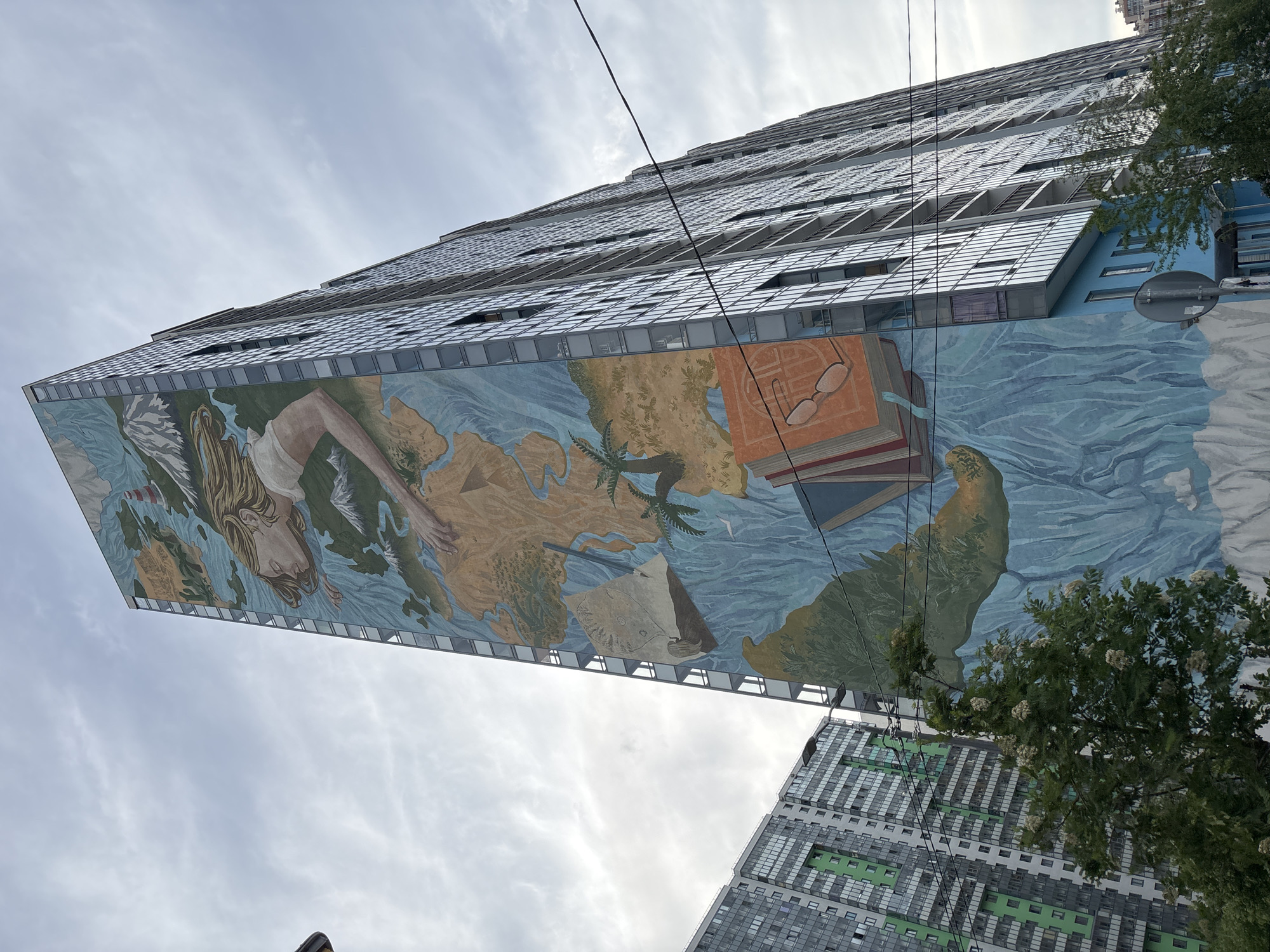 A low-angle shot captures a tall, modern building adorned with a large, vibrant mural on its facade, set against an overcast sky. The building appears to lean due to the perspective, showcasing its many reflective windows and a section of a light blue wall.
The mural depicts a complex scene: a large map-like representation of landmasses, possibly continents or islands, surrounded by swirling blue water. On the left side of the mural, the face and upper body of a woman with long, blonde hair are visible, with one arm extending across the "map." Numerous objects are integrated into the mural: a stack of books with reading glasses resting on the top book, palm trees, a white bird in flight, and a large green turtle or similar reptile partially submerged in the blue water. Further into the "ocean" area, a stylized ship or boat, resembling an old sailing vessel, is depicted.
In the lower right of the frame, another modern building, primarily grey and green, is partially visible, somewhat obscured by a leafy tree with clusters of small white flowers. Overhead, several dark power lines or cables crisscross the upper portion of the image. The lighting suggests it is daytime, under cloudy conditions. No discernible text is visible on the buildings or mural.