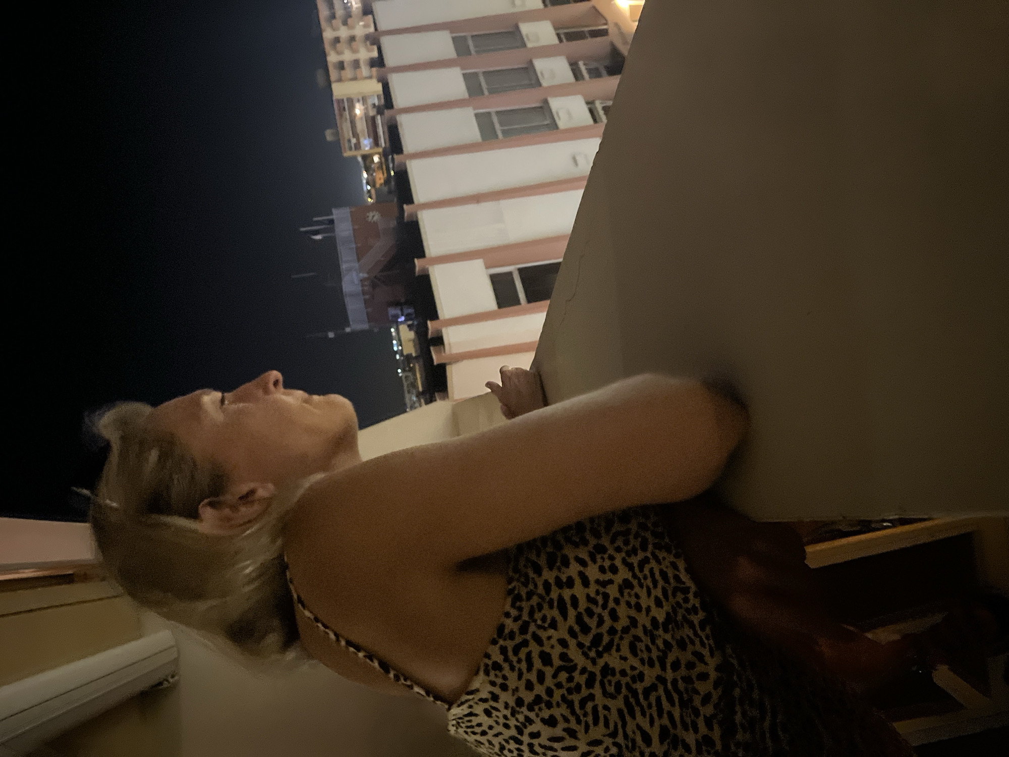A woman is standing on a balcony at night, looking out at a city skyline. She has blonde hair and is wearing a leopard-print dress. The city appears to be Palavas-les-Flots, France, based on the architecture visible in the background. The buildings are lit up, and the sky is dark. The woman's face is in profile, and she has a pensive expression. The balcony railing is in the foreground, and it is made of a light-colored material. The image is a medium shot, and the woman is the main subject.