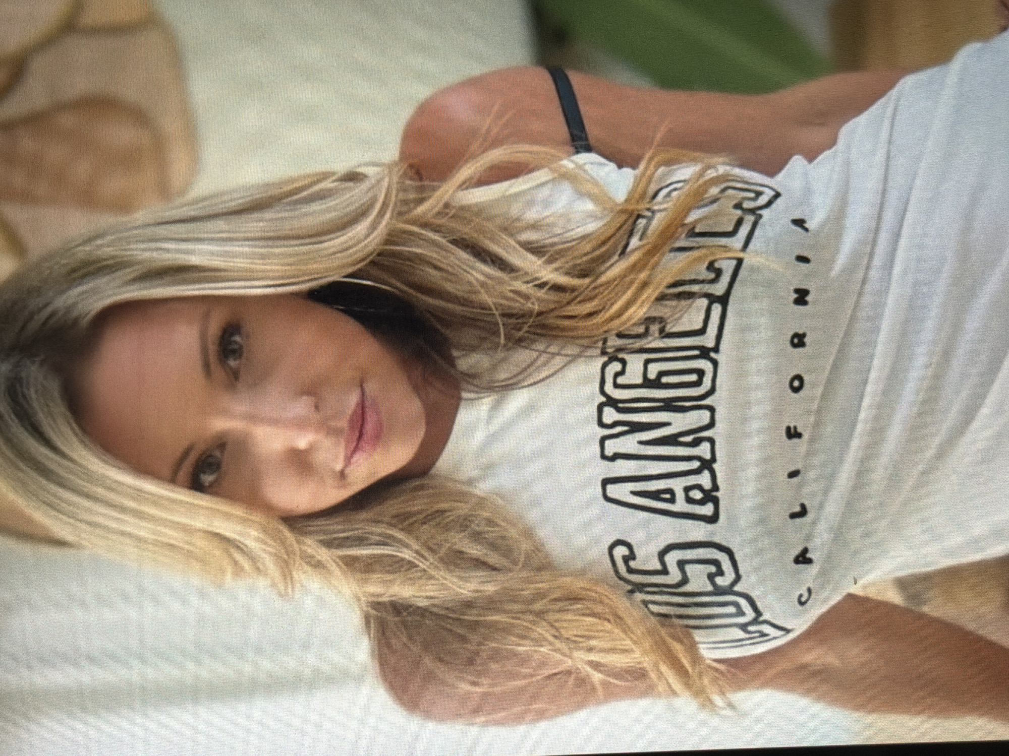 A close-up, waist-up shot features a blonde woman with long, layered hair. She is wearing a white t-shirt with "LOS ANGELES CALIFORNIA" printed on it in black lettering. The woman has light skin and is looking directly at the camera. She is wearing hoop earrings. The background is out of focus and appears to be an indoor setting with a light-colored wall and possibly some muted, earthy tones. The image does not provide any information that would allow for its location to be determined in the city of Shumen, Bulgaria.