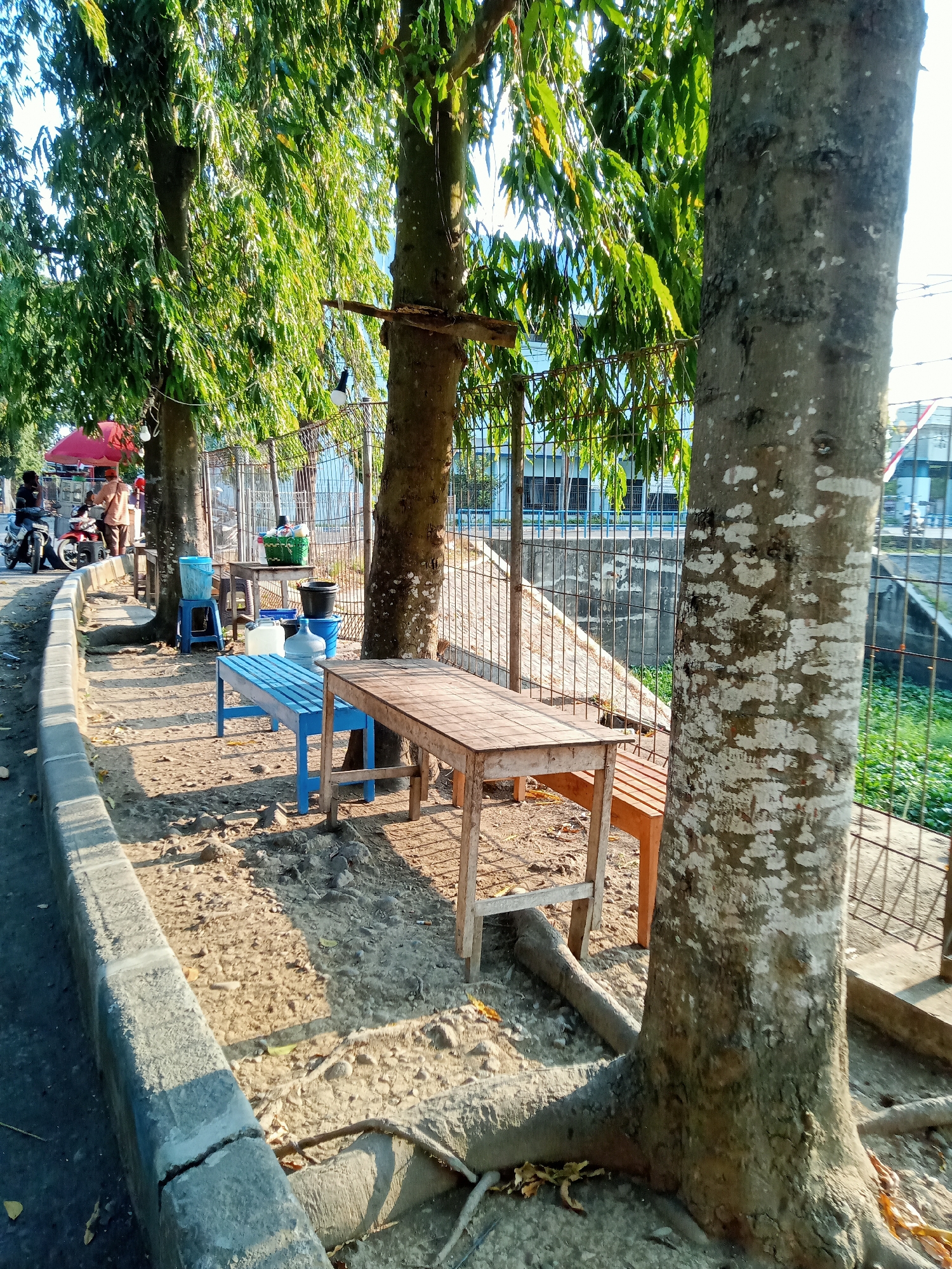 The image captures an outdoor scene in the city of Majan, Indonesia, likely along a roadside. In the foreground, a concrete curb separates a paved road from a sandy area. Several trees with lush green foliage are interspersed throughout the scene, casting dappled shadows.
In the sandy area, there are wooden tables and benches, suggesting a resting or eating area. One table is a simple wooden structure with benches on either side. Another table, also wooden, appears to be set up for serving, with a green basket containing items on top, alongside various containers, including large water jugs and buckets. A blue bench and a blue stool are also present.
Further in the background, a metal fence lines the area, beyond which a body of water with green vegetation is visible. A building with multiple windows is situated across the water. On the road to the left, two motorcycles are parked, and several people are visible, one appearing to be standing near a red umbrella, possibly indicating a small roadside stall or vendor. The overall lighting suggests it is daytime, with sunlight illuminating the scene.