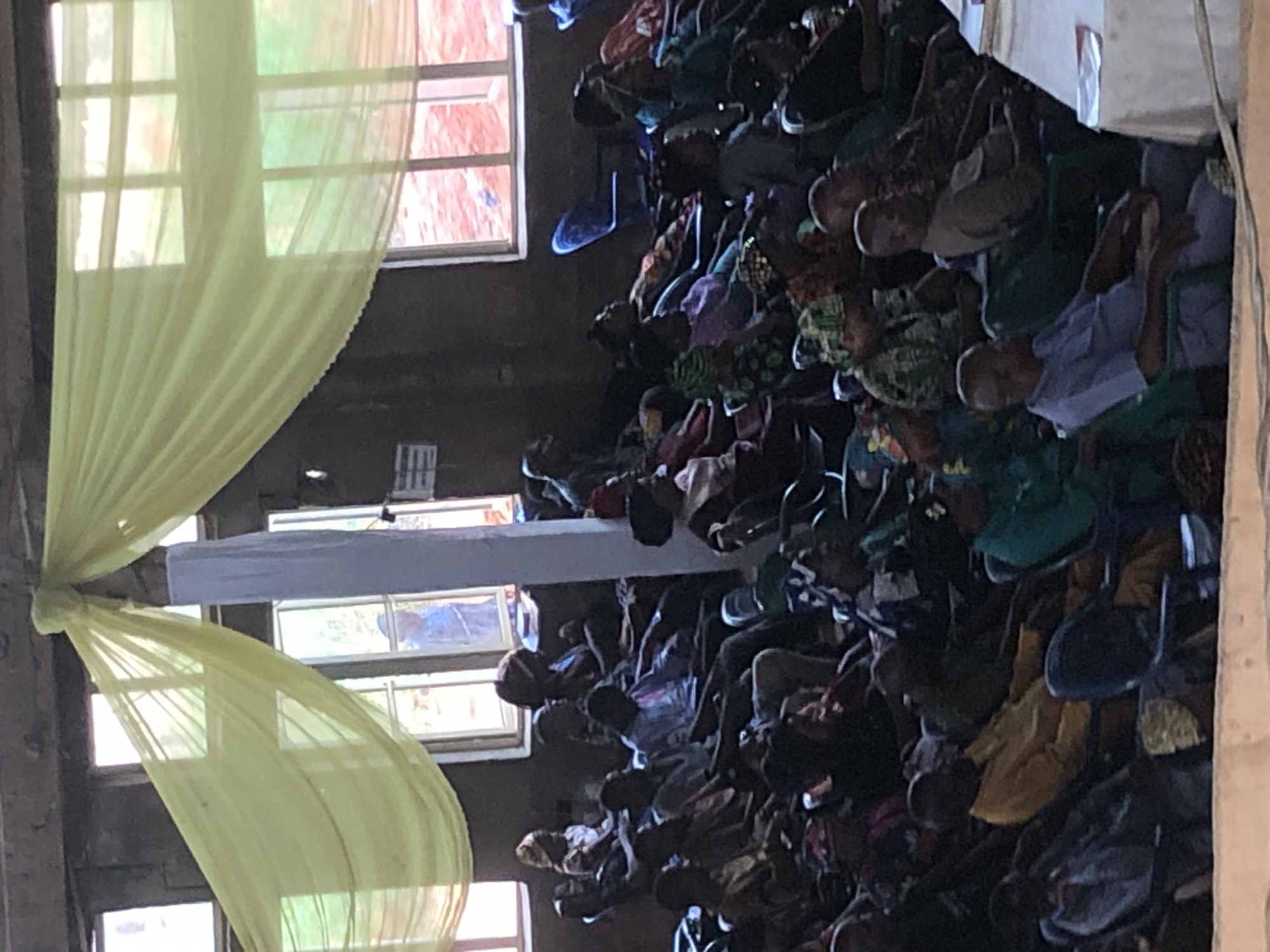 The image depicts an indoor scene, possibly a church or meeting hall, in Nteje, Nigeria. Rows of people are seated in chairs. The room is illuminated by natural light filtering through windows, some of which are adorned with pale yellow fabric drapes. A horizontal white beam or structure is visible across the top of the frame, also decorated with yellow fabric. The environment appears somewhat aged, judging by the building's structure and design.