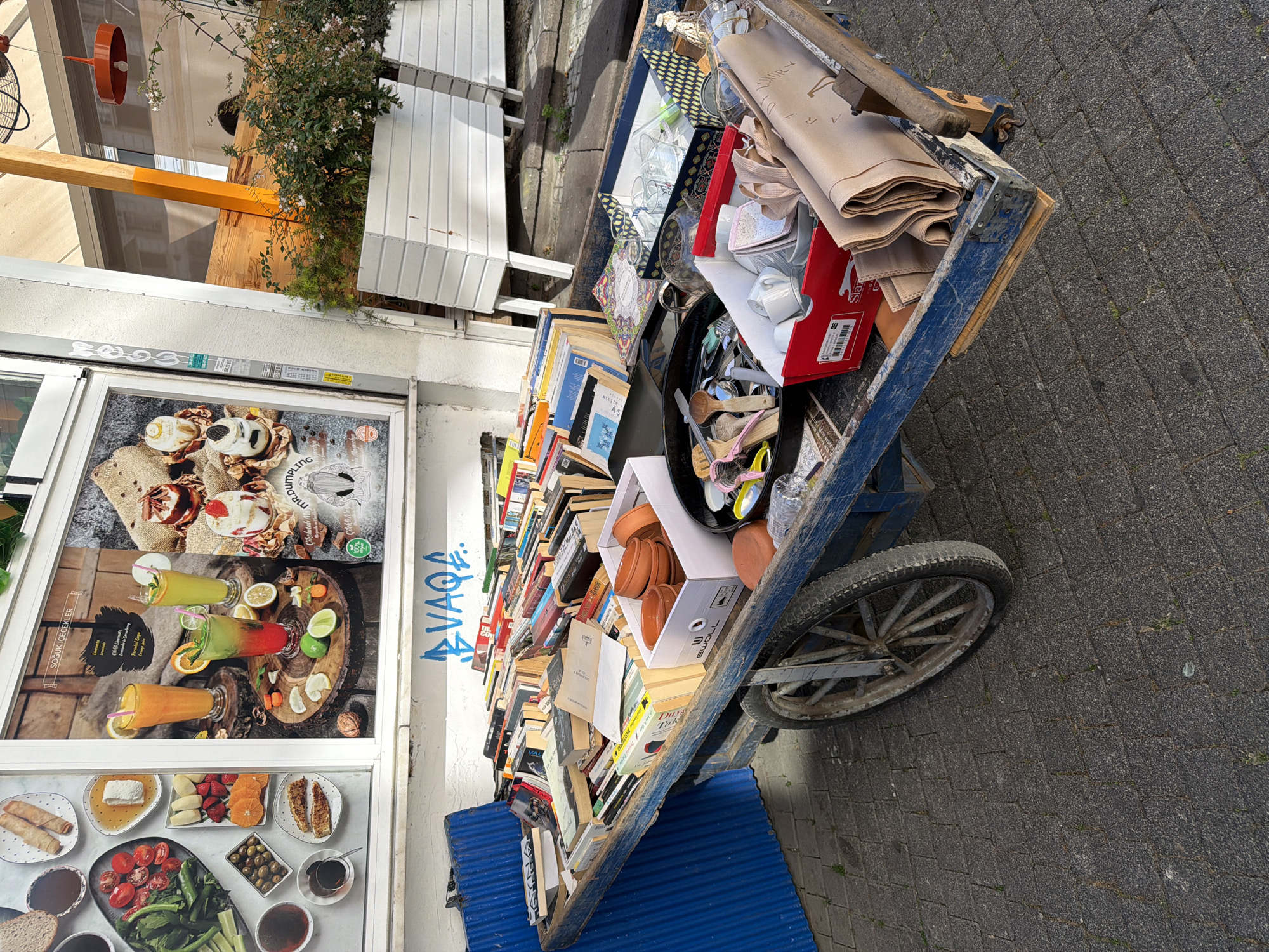 A wooden cart filled with books and assorted items is parked on a cobblestone sidewalk in Kadıköy, Turkey. The cart is laden with a variety of books, many with colorful covers, stacked haphazardly. Among the books are several boxes containing household items such as ceramic dishes, glasses, and fabrics. A black frying pan holds kitchen utensils, including wooden spoons and metal tongs. The cart is blue and shows signs of wear and tear. In the background, a storefront displays advertisements for food and beverages. To the right, a white planter filled with greenery and white flowers stands near a wooden structure. The scene appears to be outdoors, suggesting a market or a street vendor's stall.
