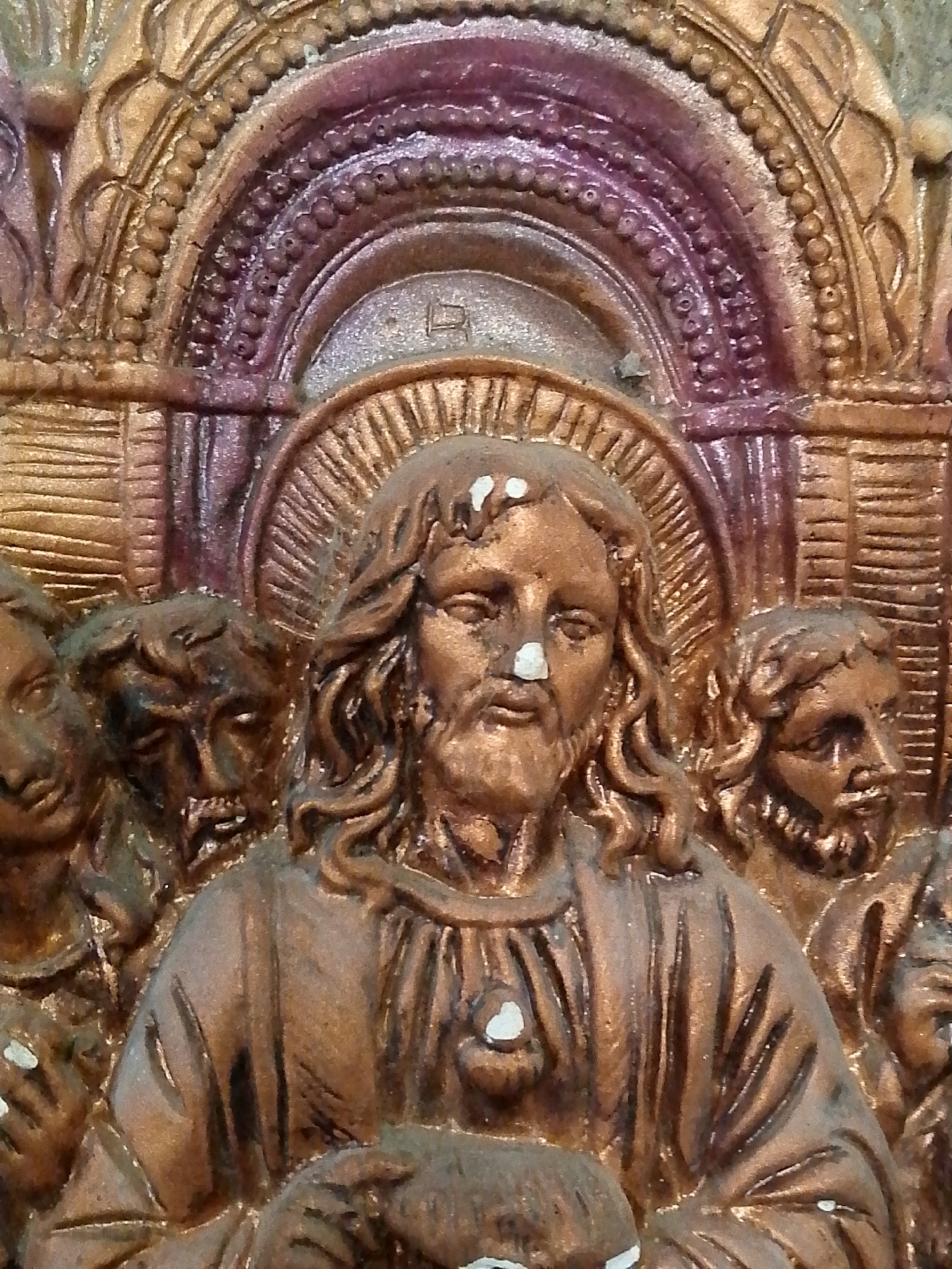 The image depicts a bronze-colored religious relief sculpture, likely representing Jesus Christ and his disciples. Christ is centrally positioned with a halo, surrounded by figures on either side. The figures appear to be engaged in the Last Supper. The background features an ornate, arched design. The sculpture is likely located in Juan Griego, Venezuela, given the request context. Specific details about the environment and placement of the sculpture within Juan Griego are not discernible from the image alone.