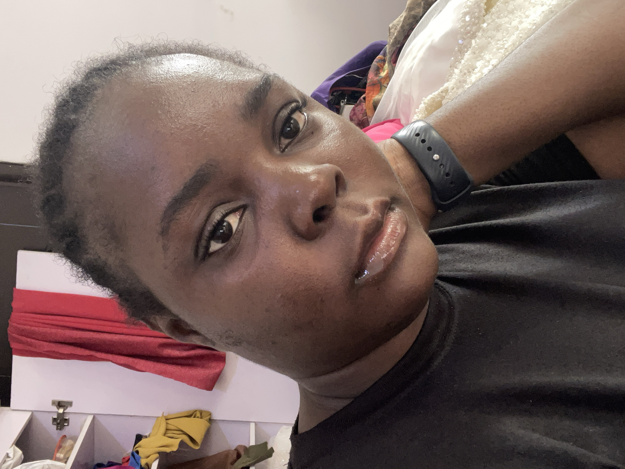 A close-up selfie shows a dark-skinned person with short, dark hair. The individual's face is slightly tilted towards the left, and their eyes are directed towards the camera. The person wears lip gloss on their lips. They are wearing a black t-shirt and a black smartwatch. The background is cluttered with items, including what appears to be a white cabinet or shelf with red fabric draped over it. Other pieces of clothing in various colors are visible in the background. The location appears to be indoors. There's no indication that this image was taken in Dushepe, Nigeria.