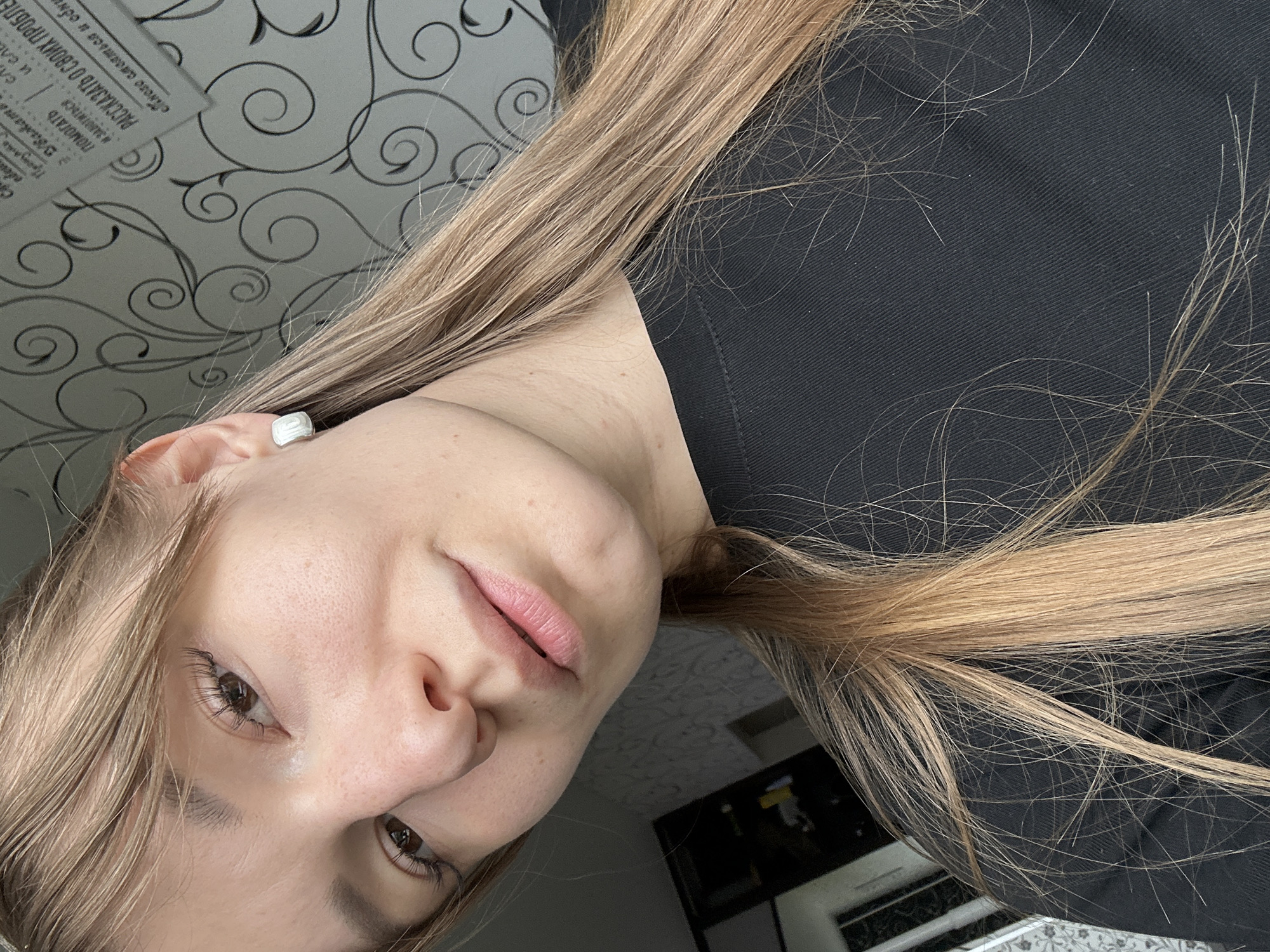Here is a description of the image:
The image is a selfie of a young woman with fair skin and light brown hair. She is wearing a black top. Her head is tilted to the side, and she is looking towards the camera. Her right ear is visible with a white square earring. The background includes a patterned wallpaper with swirls, and what appears to be a television or picture frame on the bottom right of the image. There is also a paper with some text that says "РАССКАЗАТЬ О СВОИХ ПРОБЛЕ" which translates to "TELL ABOUT YOUR PROBLEMS." The exact location within the city of Chaikovsky, Russia cannot be determined with the given information.
