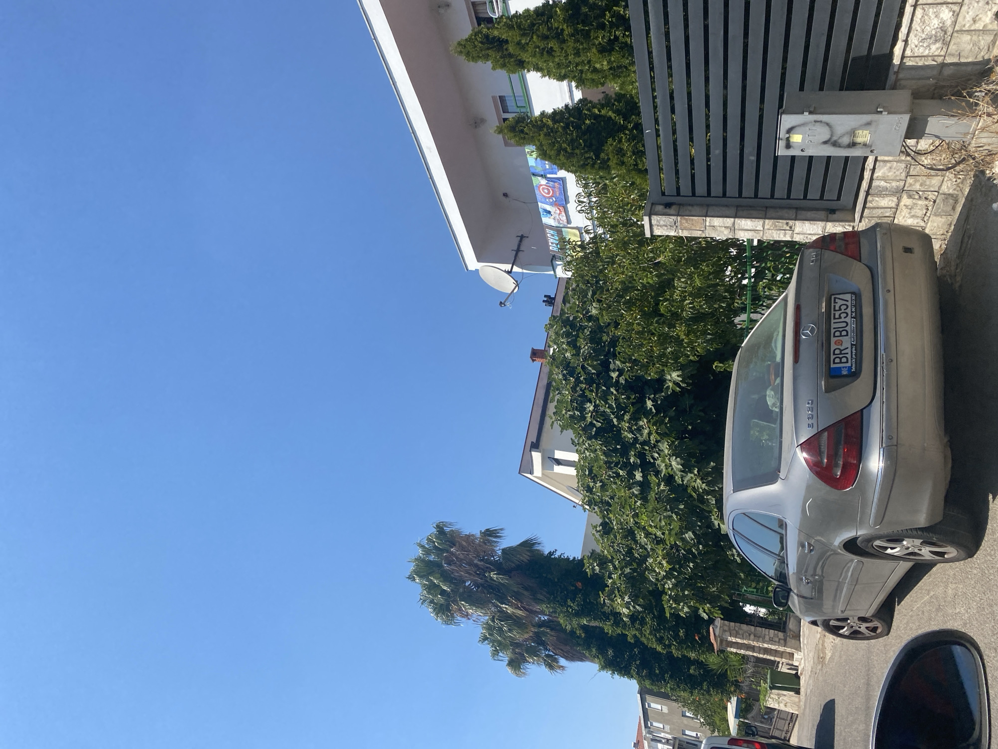 A silver Mercedes-Benz E320, license plate BR-BU557, is parked on a paved street in the city of Bar, Montenegro. The car is facing away from the camera, and its rear is visible. Behind the car is a grey stone wall topped with a black metal fence. To the left of the fence is a grey utility box with graffiti. A large green tree is behind the car, and a white building with a satellite dish and colorful towels hanging from a balcony is visible in the background. Two tall palm trees are to the left of the building. The sky is clear and blue.