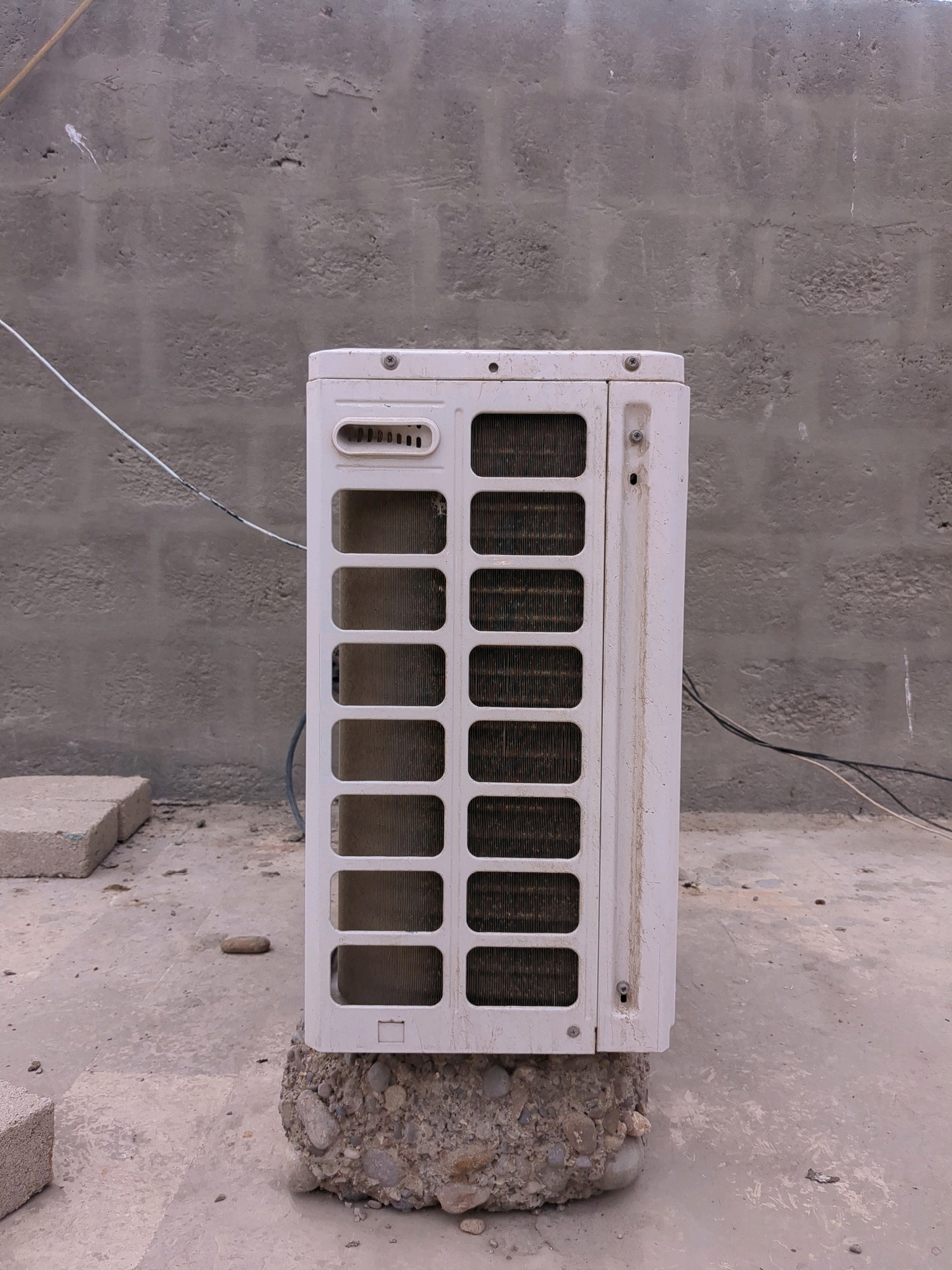 The image shows a white outdoor air conditioning unit placed on a concrete block filled with gravel and stones. The unit is positioned on a concrete surface, possibly a rooftop, with a textured gray concrete wall in the background. Several electrical wires are visible, running along the wall and near the unit. Two light-colored concrete blocks are on the left side of the frame, and a single small stone is on the concrete surface near the unit. The scene appears to be outdoors in an urban environment, likely in the city of Karachi Division, Pakistan, given the architectural style of the wall and the overall construction materials. There are no people or animals visible in the image.