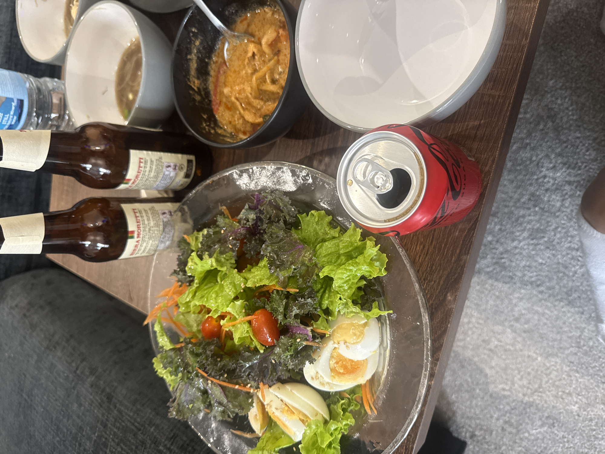 On a wooden table, two bottles of Birra Moretti beer and a can of Coca-Cola are placed next to a salad on a clear plate. The salad contains lettuce, tomatoes, shredded carrots, and sliced hard-boiled eggs. Two bowls of food, possibly curries or stews, are also present, one with a spoon. A white plate sits to the right of the Coca-Cola can. The background shows a dark-colored couch and a water bottle. The scene appears to be indoors, likely a dining area, and is set in the city of Throop, United Kingdom. There are no people clearly visible in the image.