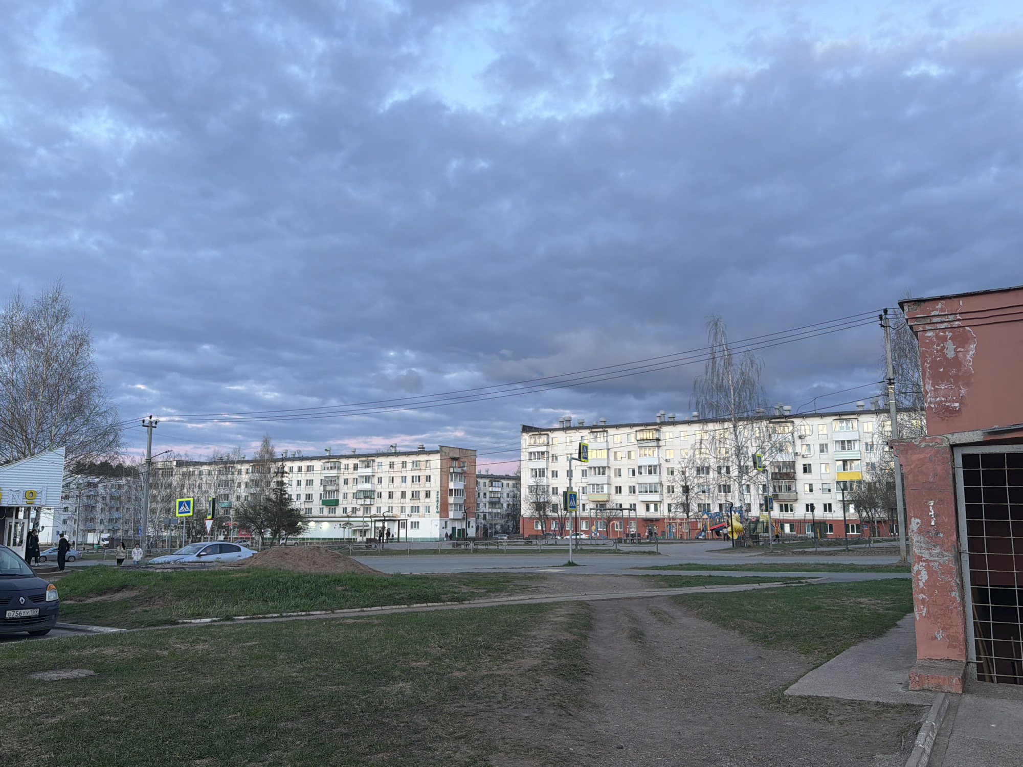 This image depicts a residential street scene in Chaikovsky, Russia, under a twilight sky. The time of day appears to be late afternoon or early evening, characterized by a soft, diffused light and a sky filled with various shades of grey and blue clouds, with hints of pink and orange indicating sunset or sunrise.
The scene is dominated by several multi-story apartment buildings, typical of Soviet-era architecture, stretching across the middle and background. These buildings are predominantly white with some green, brown, and yellow accents on balconies and window frames. In the foreground on the right, there's a reddish-pink building with visibly peeling paint and a metal grate covering an opening. On the far left, a smaller white building, possibly a shop or kiosk, features a yellow sign with a dark '9' logo.
A paved road runs horizontally across the mid-ground, with pedestrian crossings marked by blue and white signs. Grassy areas with some bare patches of earth and a dirt path are visible in the foreground. Leafless trees, likely deciduous and indicating early spring or late autumn, are scattered throughout. Overhead utility lines and poles crisscross the upper portion of the image.
Several individuals are present, mostly in the middle ground near the buildings and road, either walking or standing. A dark-colored car is partially visible on the far left, with its license plate "0756TY 159" clearly readable, which is a regional code for Perm Krai, Russia. Another light-colored car is visible near a mound of sand or dirt in the center-left. The overall atmosphere is quiet and routine.