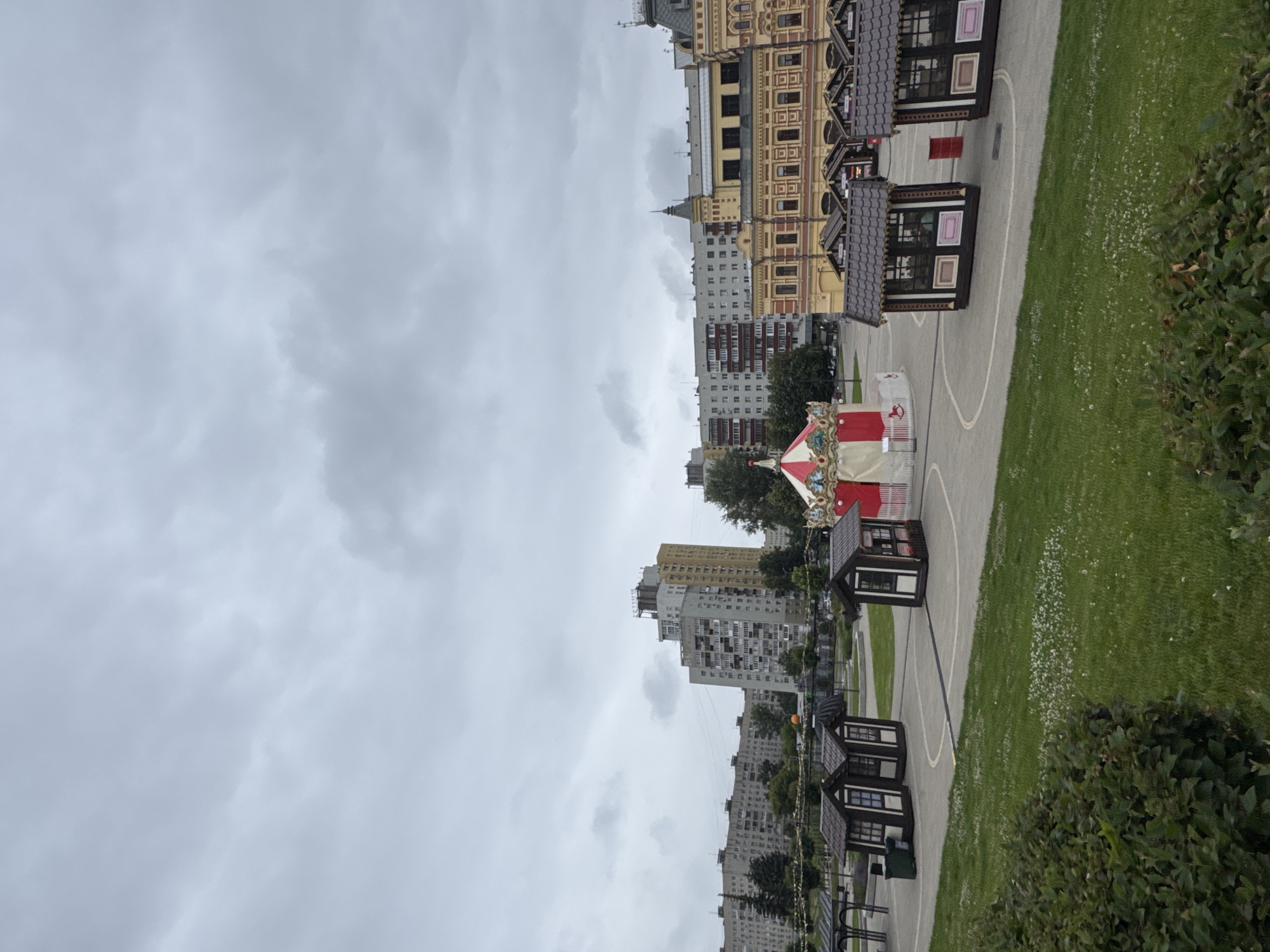 The image captures a cloudy day in Nizhny Novgorod, Russia. The foreground features a well-maintained green lawn bordered by dense green bushes. In the middle ground, a paved circular area hosts a small, white and red striped carousel, two small, dark brown wooden kiosks with brown roofs, and another similar kiosk slightly further away, to the right. Another kiosk is visible in the distance, to the far left, partially obscured by a metal structure. Behind the carousel and kiosks, a row of trees lines the background. Further back, a series of multi-story residential buildings, characteristic of Soviet-era architecture, stretch across the horizon. To the right of these buildings, a distinct, ornate building with yellow and red facades and intricate detailing stands out. The sky is overcast with thick, gray clouds, suggesting an impending or recent rainfall.