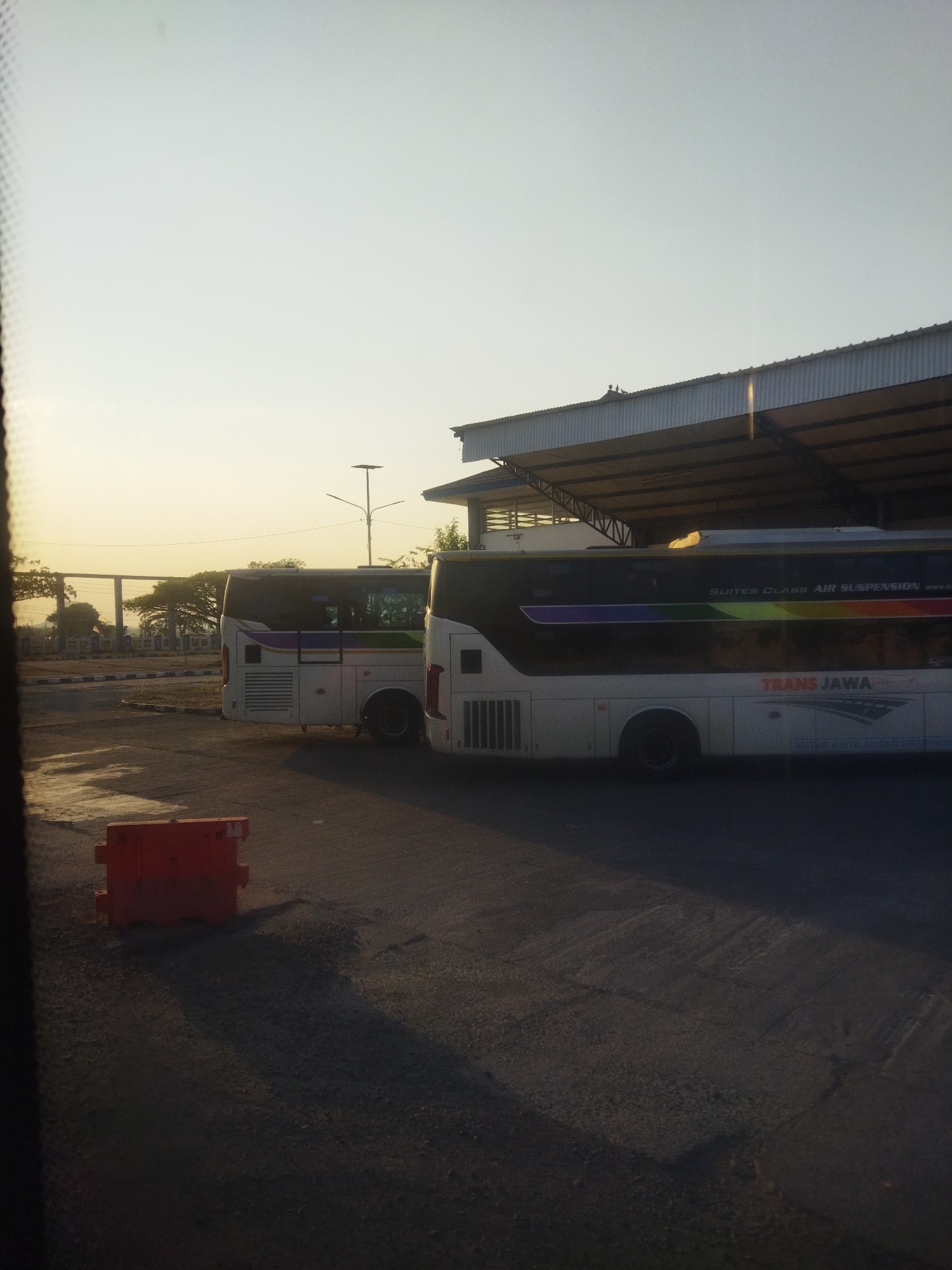 In Kaliancar, Indonesia, two white buses are parked at a bus station under a covered shelter. The bus on the right, labeled "TRANS JAWA," has "SUITES CLASS AIR SUSPENSION" written on its side, along with a rainbow-colored stripe. The bus on the left is partially visible and has a purple, green, and yellow stripe. An orange traffic barrier is in the foreground. The sky is clear and pale, suggesting it is either early morning or late afternoon. The ground is paved, and shadows are cast across it. The overall scene depicts a quiet moment at a transportation hub.