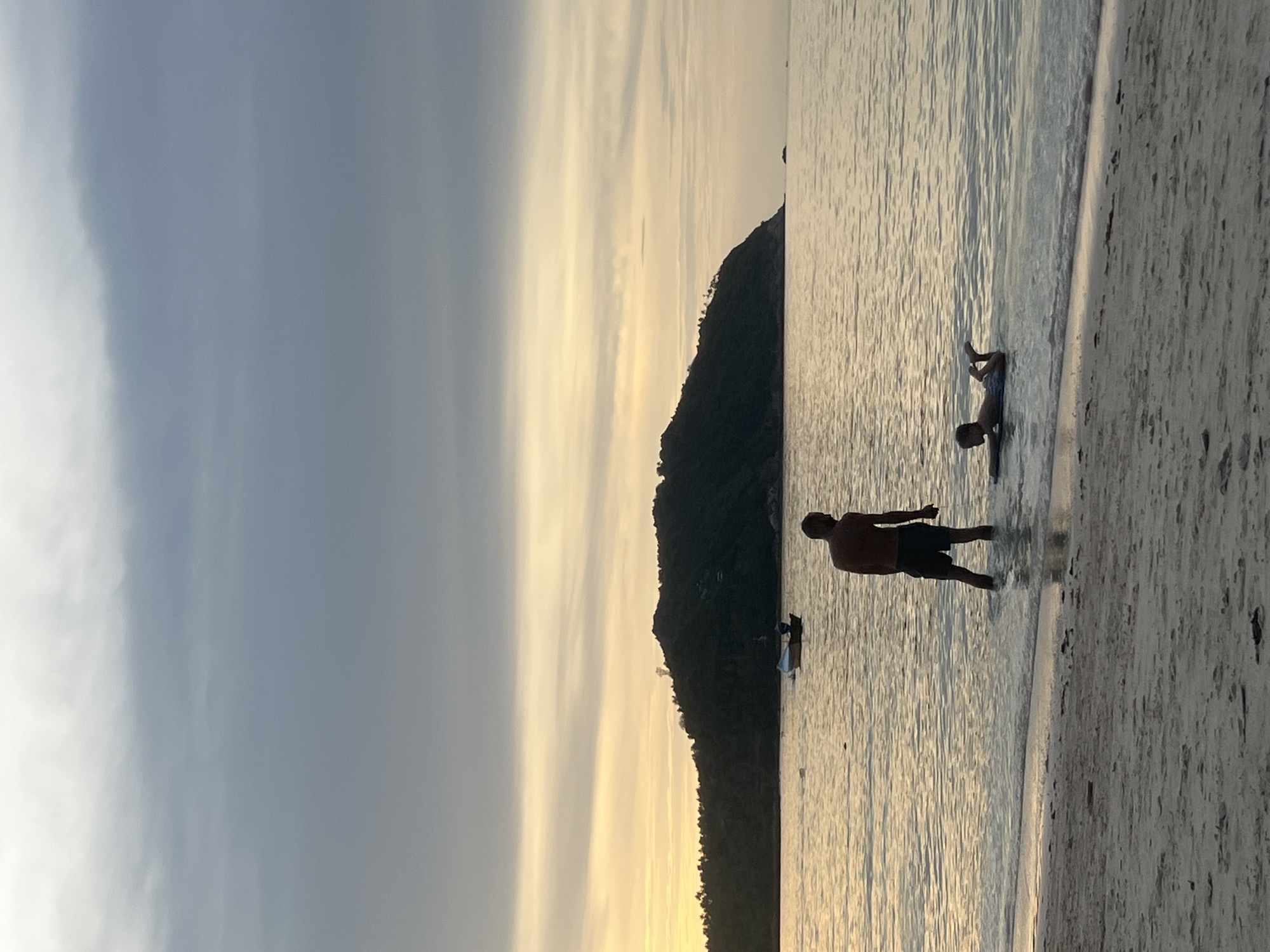 Two individuals are in the shallow water of a beach at sunset on Ko Pha-ngan District, Thailand. One person, an adult male, stands in the water, facing away from the camera, wearing dark shorts. Another person, a child, lies on their stomach on a bodyboard, also in the shallow water, facing left. The sun is setting, casting a warm glow on the water and the sky, which is filled with grey and yellow clouds. In the background, a dark, tree-covered hill rises from the sea. A small fishing boat is visible in the water near the hill. The foreground consists of a sandy beach.