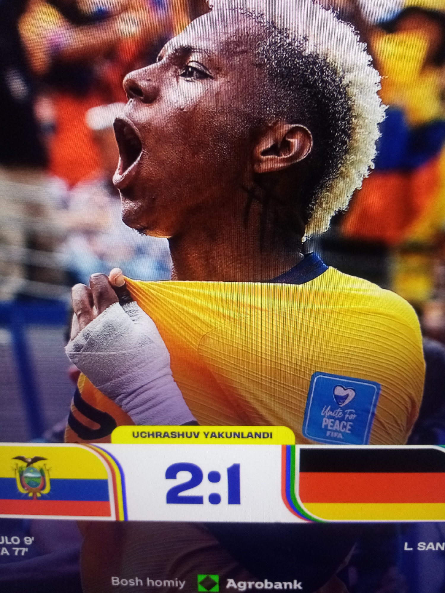 This media file captures a close-up of a soccer player with distinctive bleached-blonde hair, wearing a yellow jersey. The player is seen with their mouth wide open, seemingly in an expression of intense emotion, possibly celebration or exertion. Their hand, wrapped in white bandages, is pulling at the fabric of their jersey. On the jersey, a blue square patch with the text "Unite For PEACE FIFA" and a heart symbol is visible.
Below the player, a score graphic displays "2:1" between the flags of Ecuador and Germany. The flags are positioned to the left and right of the score, respectively. Additional text elements appear at the bottom of the frame, including what seems to be a time stamp ("JLO 9'", "A 77'"), the word "Agrobank" with a green logo, and "Bosh homiy". The phrase "UCHRASHUV YAKUNLANDI" is prominently displayed in a yellow banner, suggesting the end of a match. The overall impression is a moment captured during or immediately after a competitive soccer match, with the player's raw emotion and the score graphic providing key contextual details. The setting appears to be a stadium, with a blurred background of spectators.