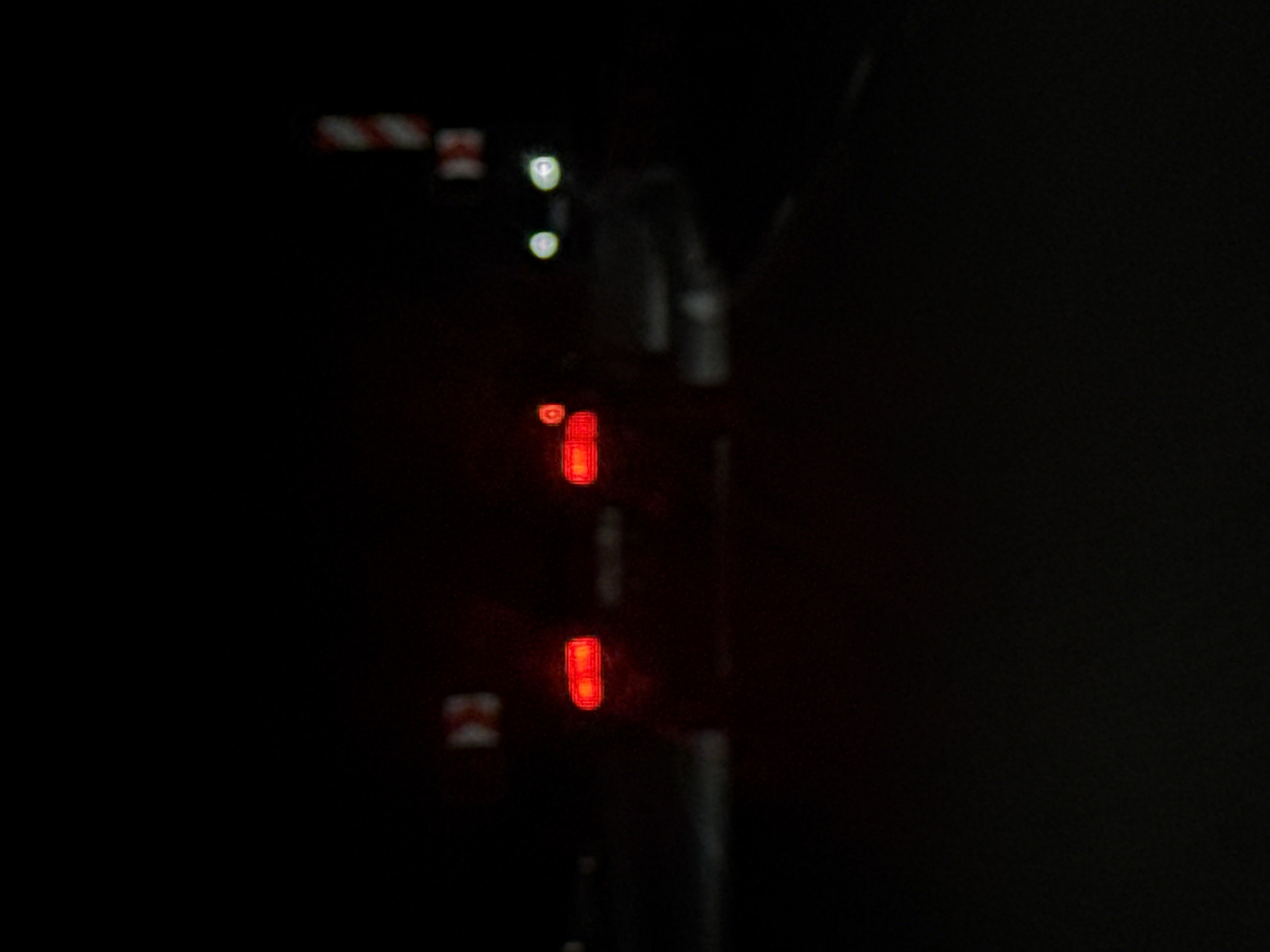 Two red lights are stacked vertically on a gray pole in the darkness. A red and white chevron sign is to the left of the top light. Two fuzzy white lights are near the top of the pole. Another red and white chevron sign is to the left of the bottom light. The location is Narechen, Bulgaria.