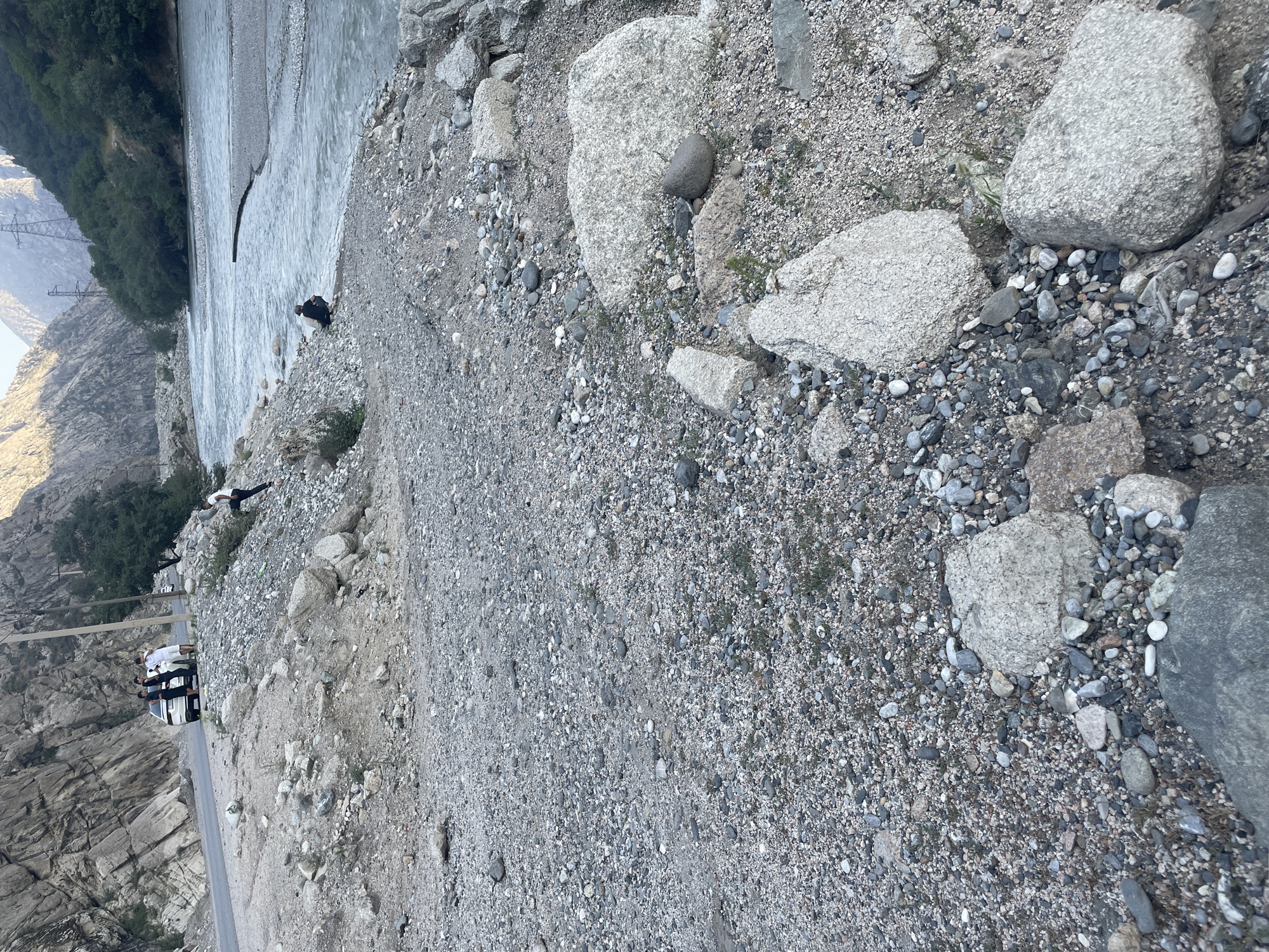 The image depicts a scene in a mountainous region, likely near Khodzha-Obigarm, Tajikistan. A paved road runs along the left side of the image, with a steep, rocky mountainside rising behind it. Several people are gathered near a white SUV parked on the side of the road. To the right, a wide, flowing river is visible, bordered by a gravelly embankment. Another person is seen crouching by the riverbank. The environment is rugged and natural, with scattered rocks and sparse vegetation on the slopes and along the river. The overall impression is of a remote, scenic location with a few individuals enjoying the outdoors.