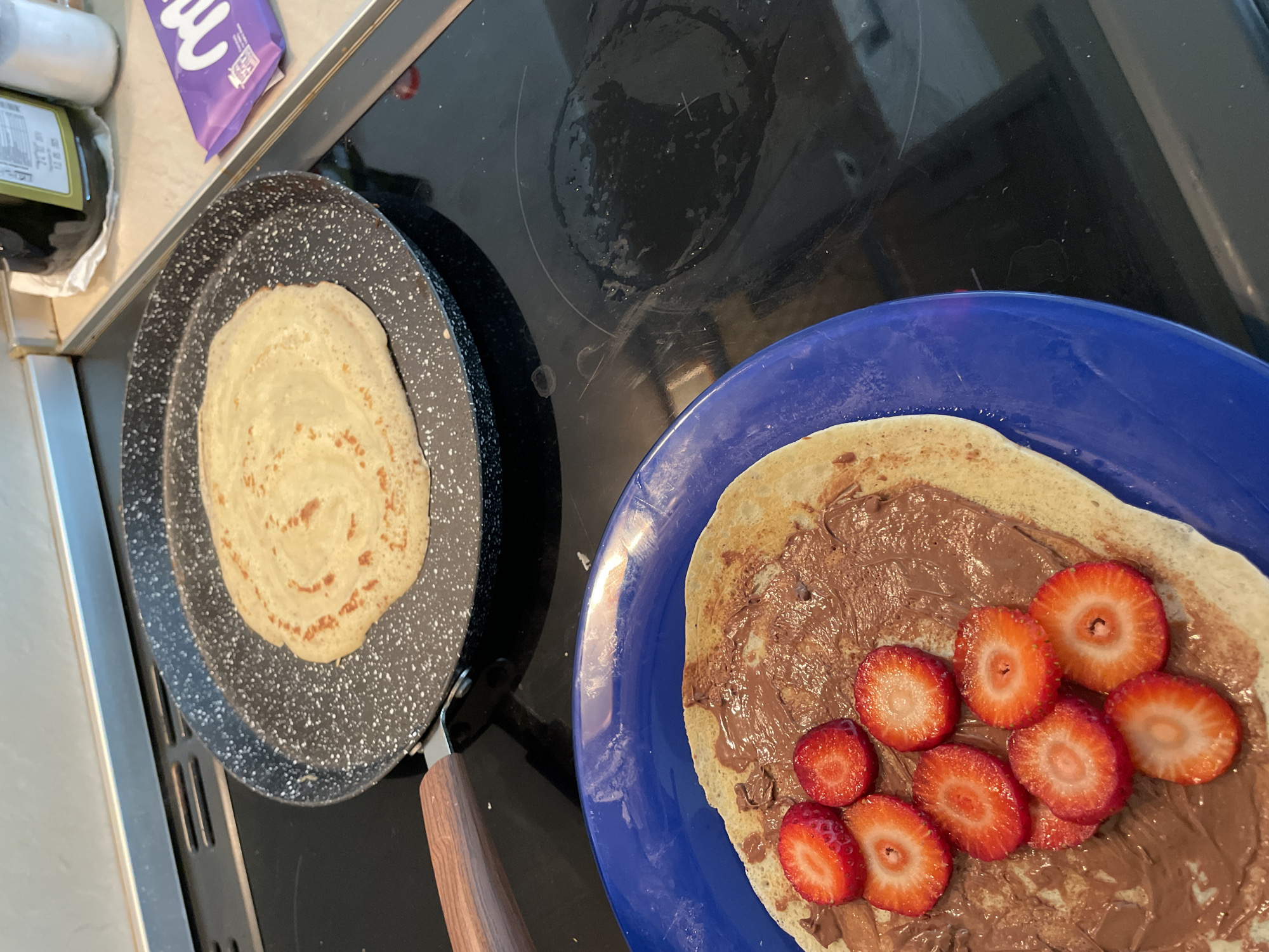 This media file captures a close-up, overhead view of a cooking scene in a kitchen, likely in Zlatograd, Bulgaria. The central focus is on two flatbreads or crepes.
On the left, a round, pale yellow flatbread is cooking in a speckled black non-stick frying pan with a wooden handle, resting on a black electric or induction stovetop. The flatbread shows some light browning, indicating it's still being prepared.
To the right, a finished flatbread is presented on a round, dark blue plate. This flatbread is generously spread with a thick layer of chocolate hazelnut spread, resembling Nutella. On top of the chocolate spread are several bright red, sliced fresh strawberries, arranged in a scattered pattern.
In the upper left background, part of a kitchen counter is visible with a dark green bottle, possibly olive oil, and a purple wrapper of a "Milka" chocolate bar. The black stovetop surface, which occupies a significant portion of the image, shows some smudges and reflections.
The image depicts the preparation and presentation of a sweet meal, possibly breakfast or a snack, featuring crepes with chocolate spread and strawberries. The overall lighting suggests daytime. No people are visible.
